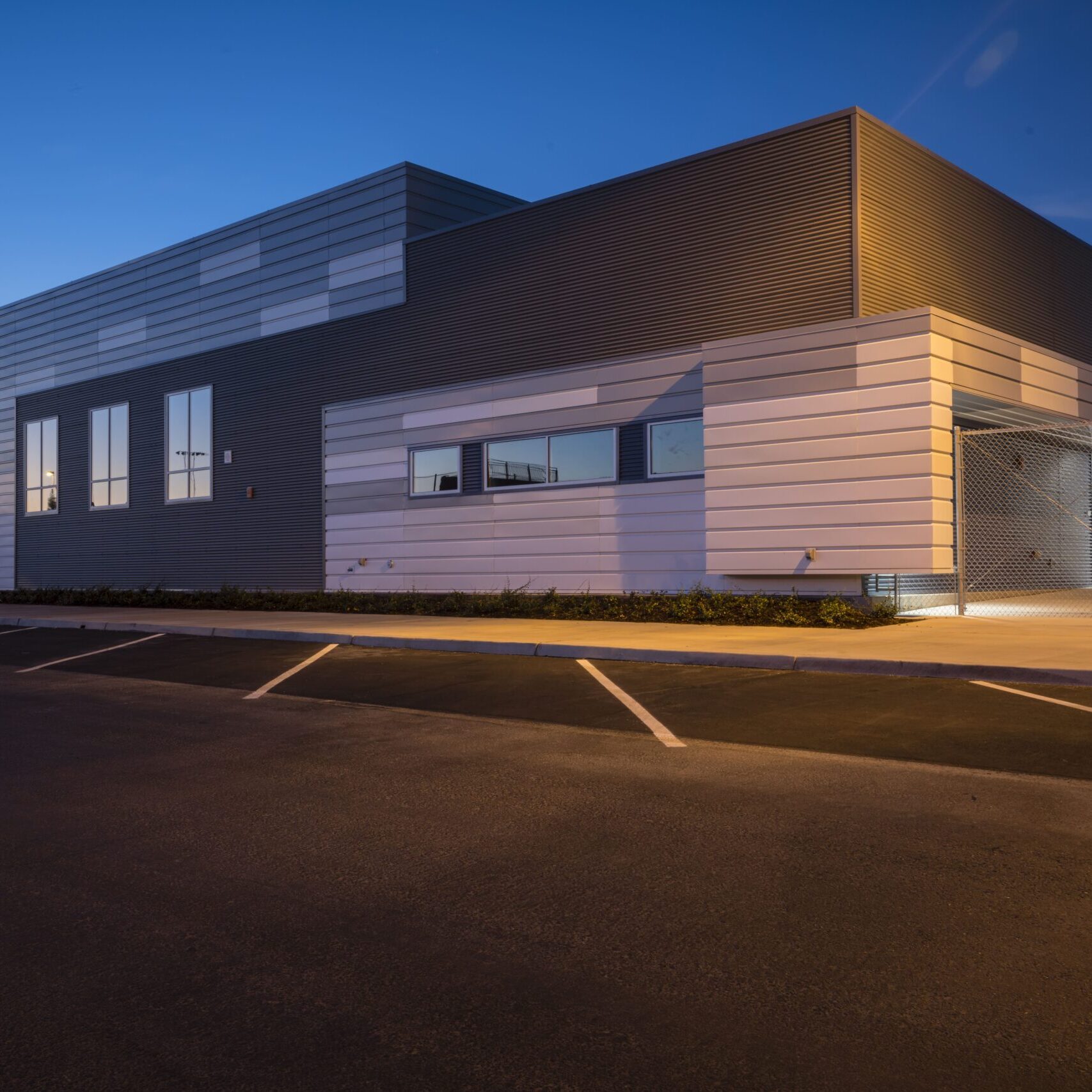 An exterior shot of a modern building at dusk with gray and white paneling. There are three large windows on the facade. The parking lot is in the foreground, and red light trails from a passing vehicle are visible on the pavement. A street lamp is lit on the right.