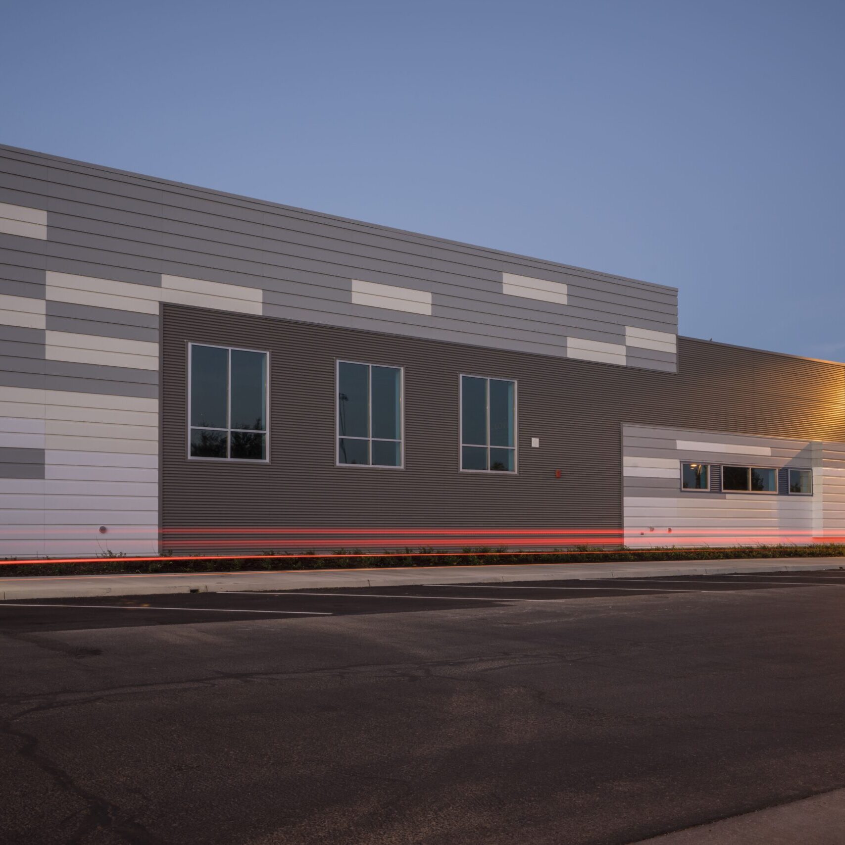 An exterior shot of a modern building at dusk with gray and white paneling. There are three large windows on the facade. The parking lot is in the foreground, and red light trails from a passing vehicle are visible on the pavement. A street lamp is lit on the right.
