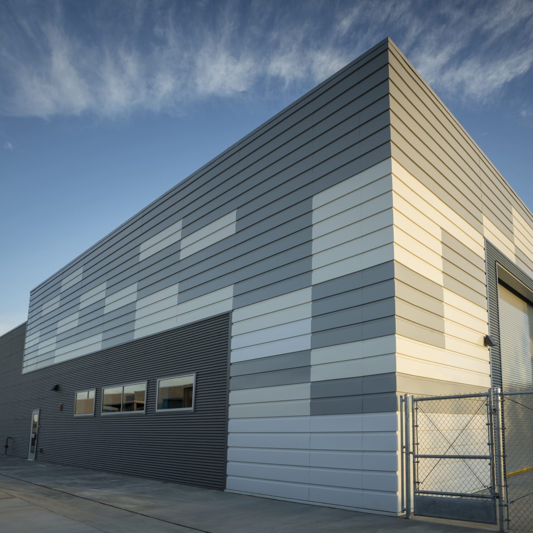 An exterior, low-angle shot of a contemporary building with gray corrugated and white paneled siding. The building is designed with a mix of horizontal and vertical panels. The sky is blue with scattered clouds, and the building is surrounded by a paved area and a chain-link fence.