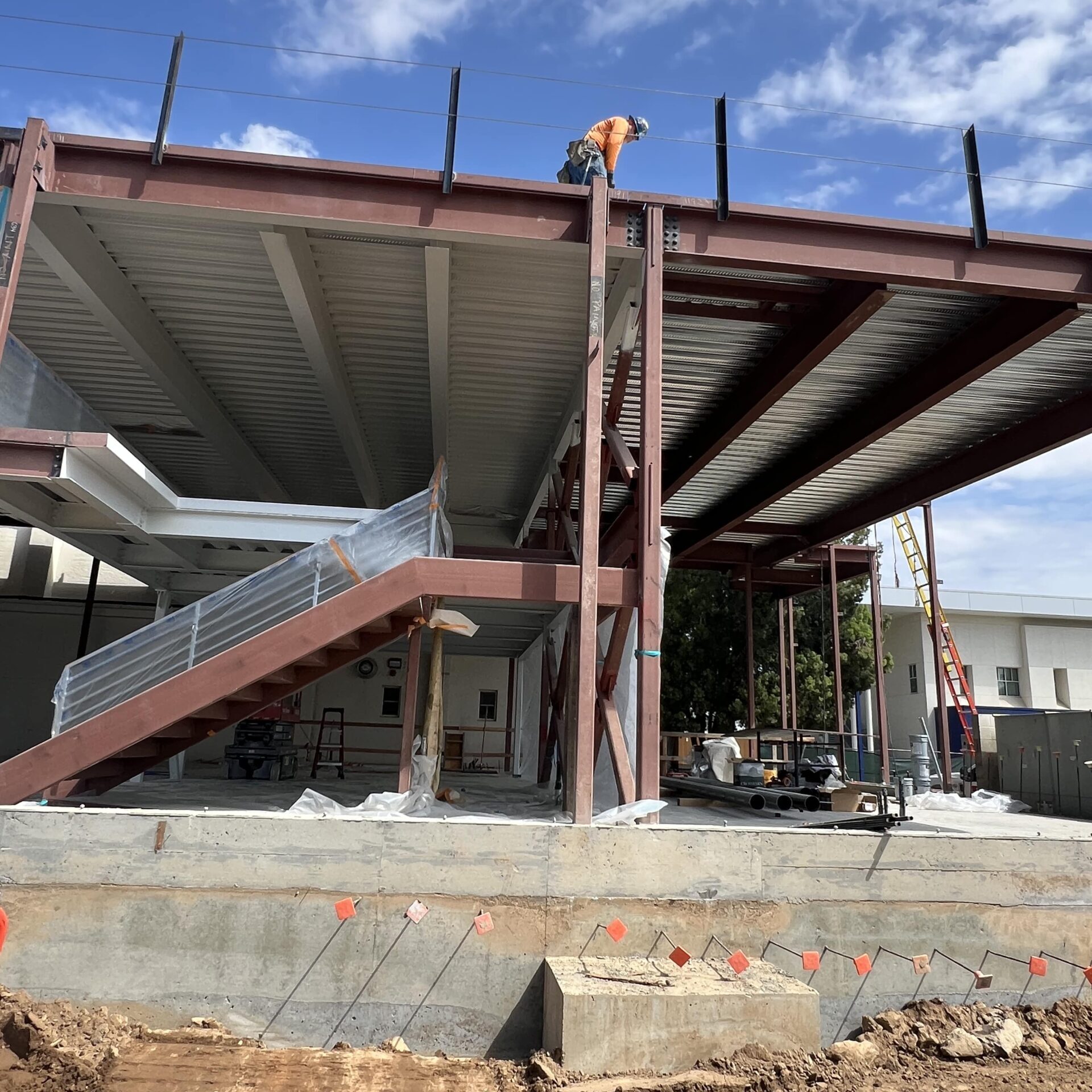 A construction worker in an orange shirt and helmet stands on a partially-built second floor of a building. The structure is made of brown steel beams. In the foreground, there's a concrete foundation and a mound of dirt.