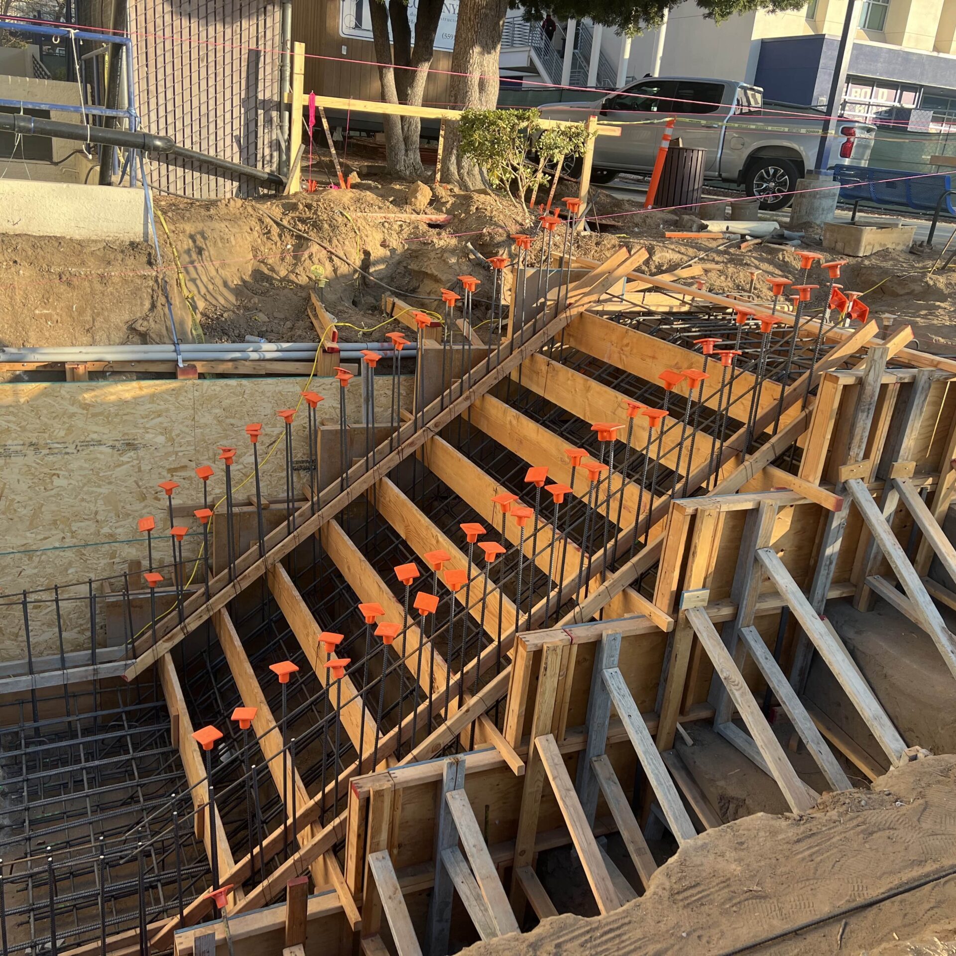 A close-up shot of a wooden staircase frame under construction in a dug-out area. The staircase has a metal rebar grid with orange caps.