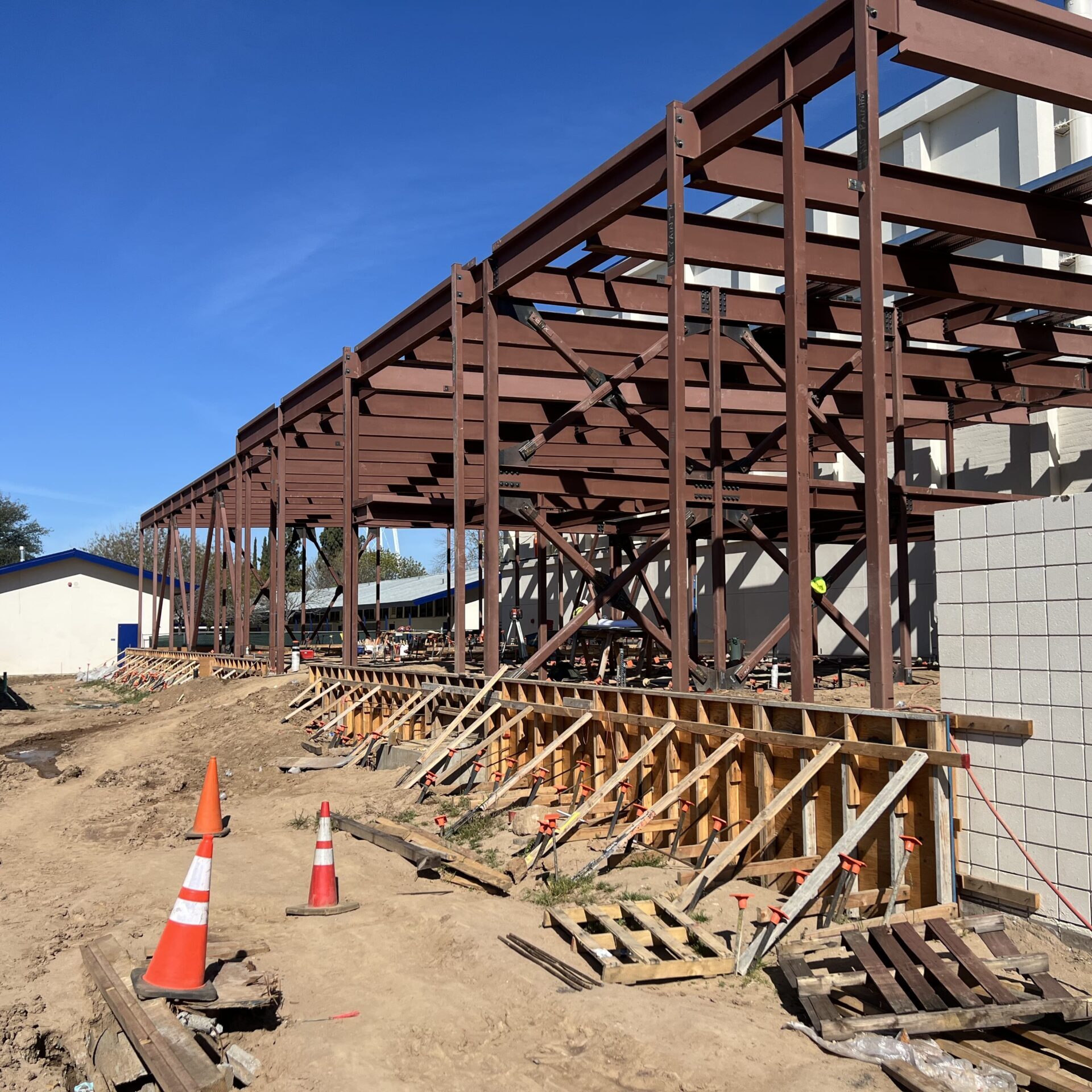 A wide shot of the steel frame of a building. The frame is brown with multiple horizontal and vertical beams. The ground is dirt, with wooden forms and orange cones.