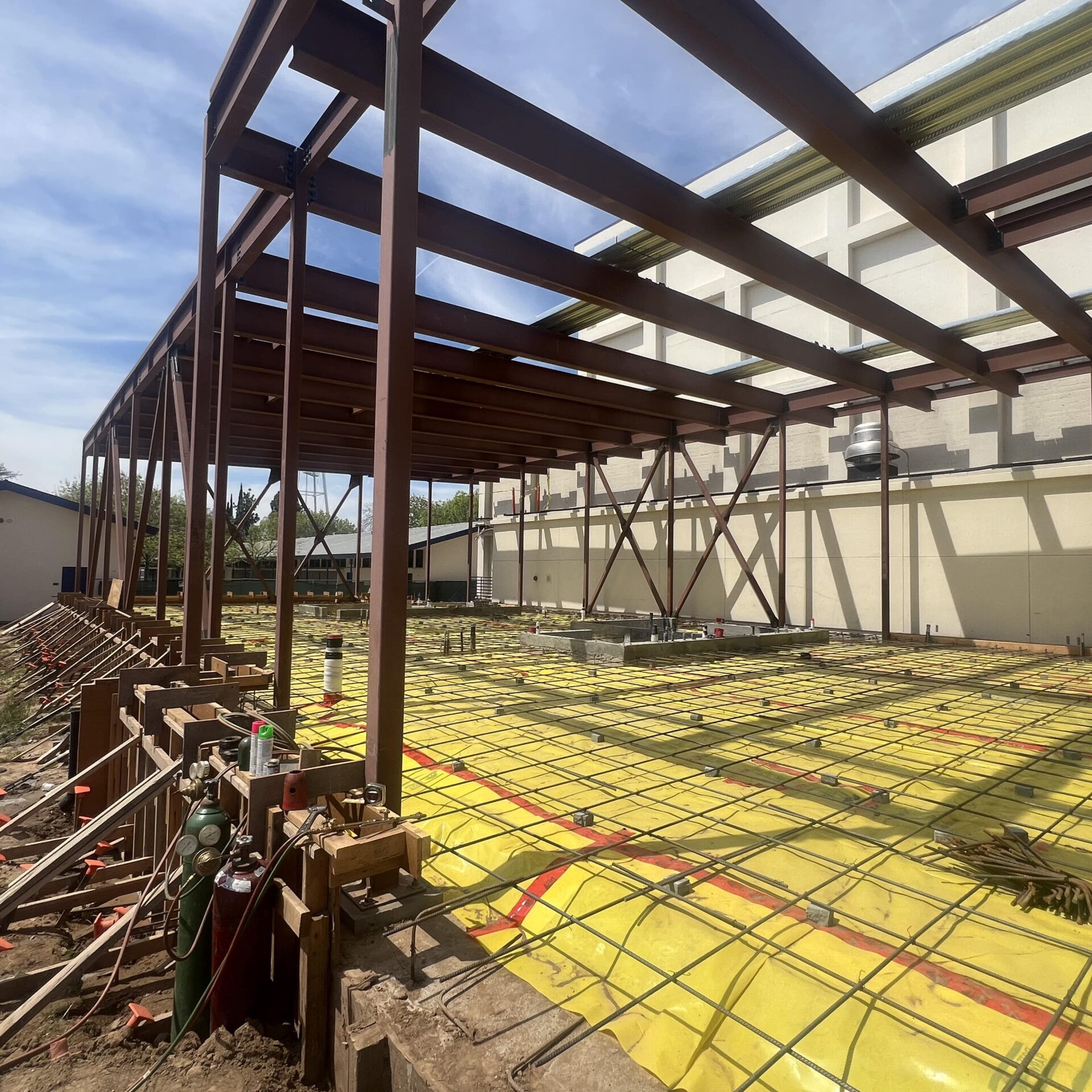 An eye-level view of the building's ground floor, which is covered in a yellow tarp and a grid of metal rebar. The brown steel frame of the building is visible around the perimeter.