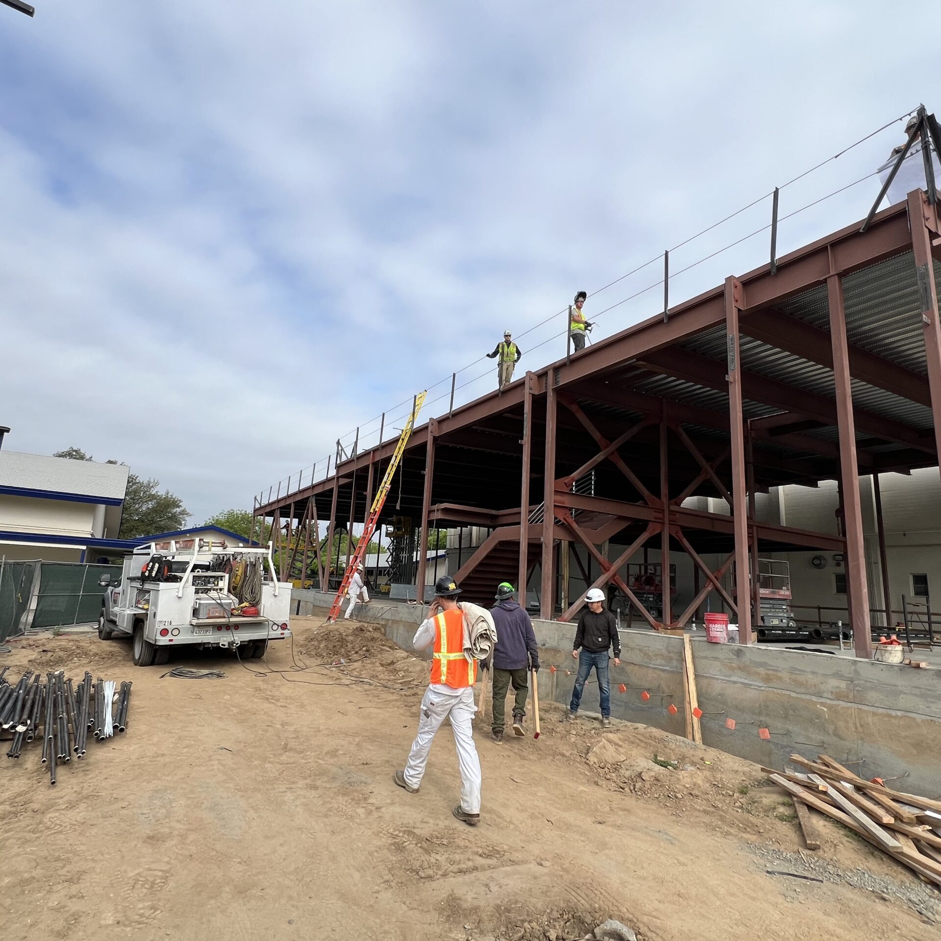 A group of three construction workers stands near the base of the building's steel frame. A white construction truck is parked nearby. Two other workers can be seen on the second floor, which has a temporary safety railing.