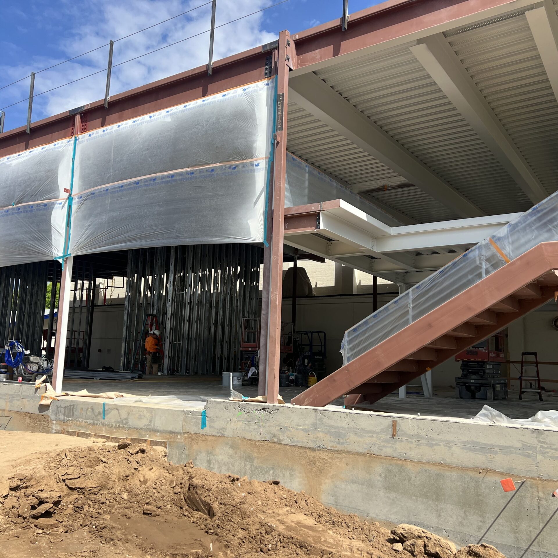 A large brown steel building frame with a staircase on the right. The second-story section is partially covered in a large sheet of white plastic. The area beneath the second story is filled with vertical metal studs. The ground is dug up and muddy.