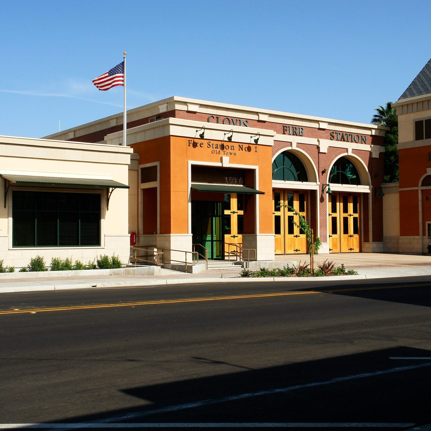 A wide, low-angle shot of the completed Clovis Fire Station No. 1 from across the street. The building is a combination of light-colored stucco and red brick, featuring three garage bays with arched doorways. An American flag flies from a pole in front of the building. The address, "633 Pollasky Ave," is visible on the left side of the building.