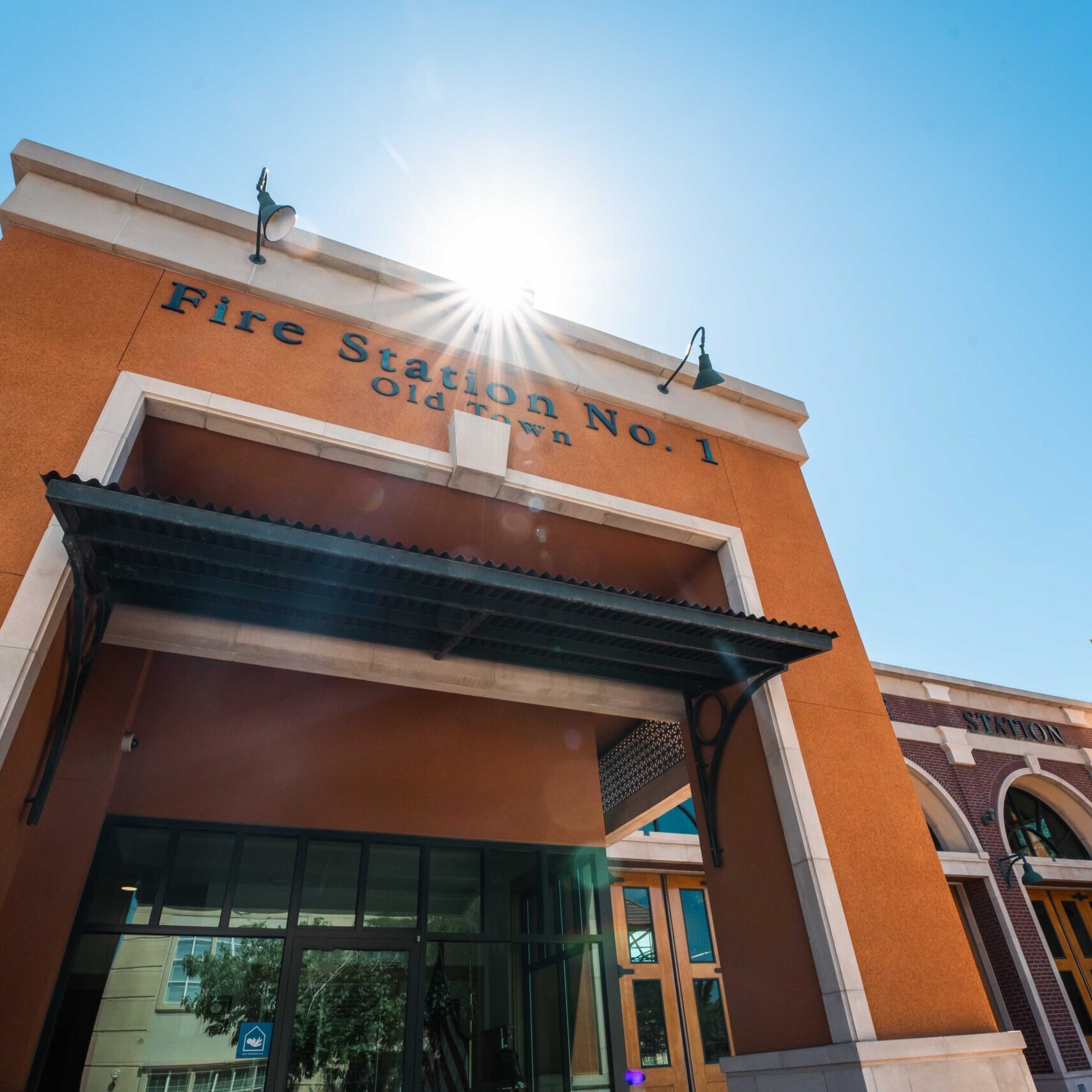 A low-angle, sunlit view of the front of the completed Clovis Fire Station No. 1. The photo shows the orange stucco facade of the building, with the words "Fire Station No. 1 Old Town" written above the main entrance. The sun is directly above, creating a lens flare effect. Part of the adjacent red brick section and the tower are visible to the right.