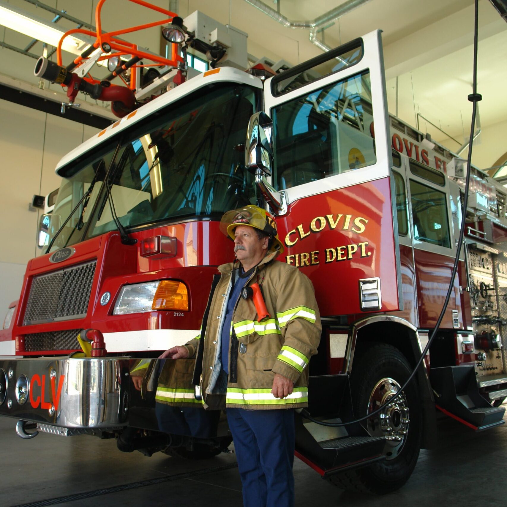 A firefighter in a full uniform stands in front of a red fire truck inside the fire station's garage bay. The truck has "CLOVIS FIRE DEPT." written on the side. The fire station's high ceiling and large arched doorways are visible in the background.