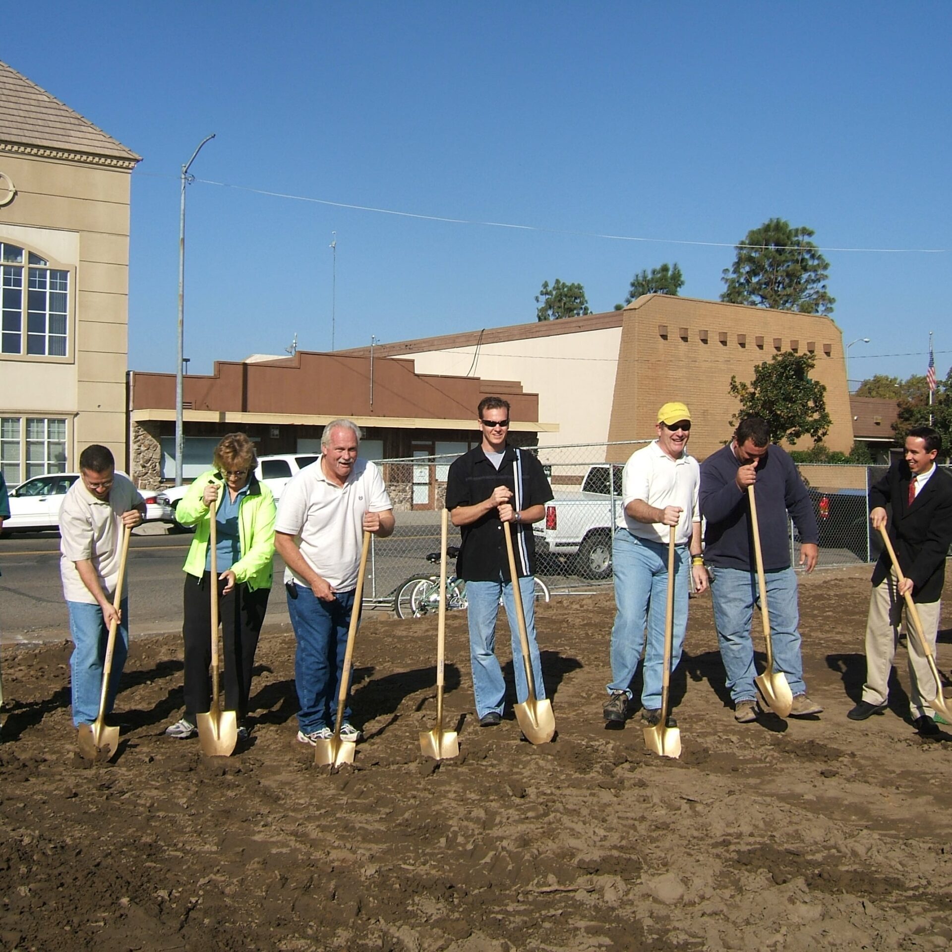 A group of people, including men and women, standing in a line on a dirt lot holding shovels for a groundbreaking ceremony. A building is visible in the background, along with a blue tent and chairs to the right.