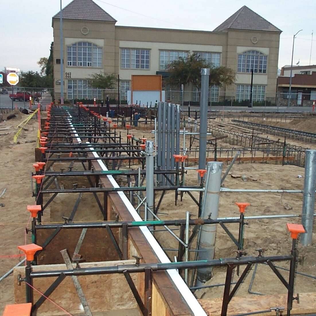 A wide shot of the early stages of construction on a large, sandy lot. The ground has been excavated and a complex grid of steel rebar and wooden forms for the foundation is visible. A multi-story building can be seen in the background.