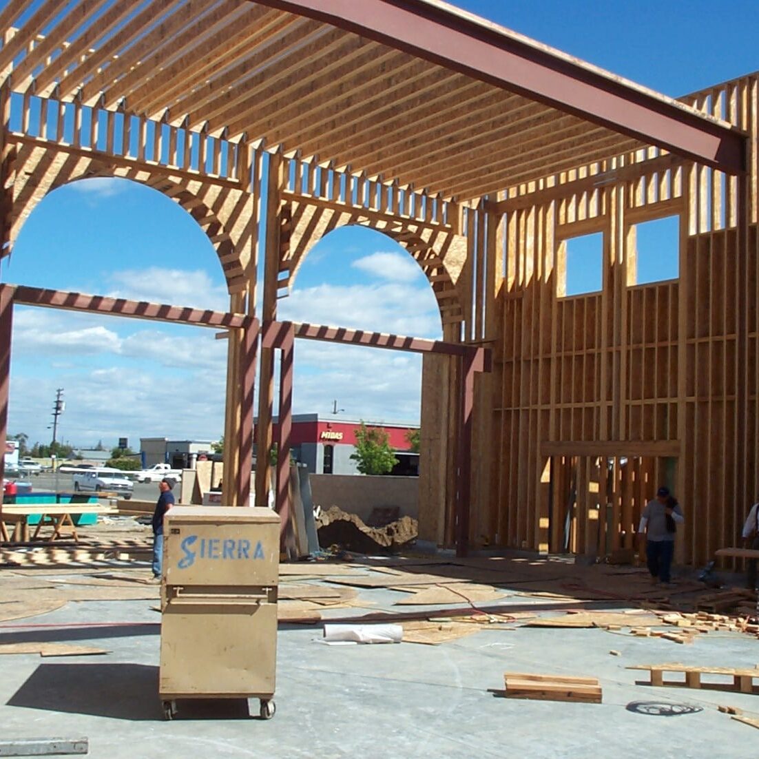 The interior of the fire station under construction. The building's frame is visible, showing tall wooden beams and an open structure with high ceilings and large arched openings. People are working in the background and a yellow toolbox is in the foreground.