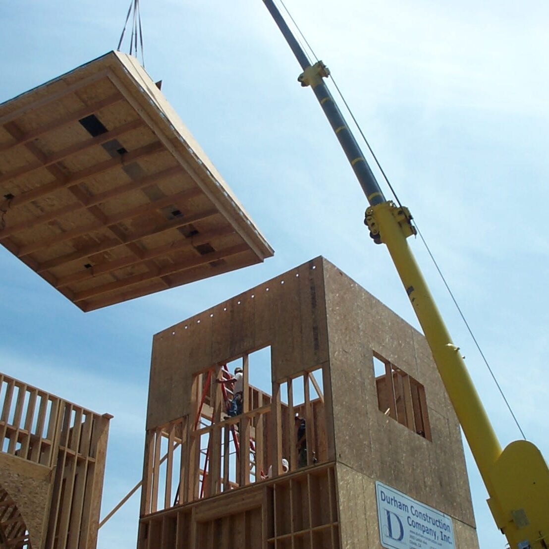 A large crane lifts a pre-fabricated wooden wall or roof section into place on the fire station's partially framed tower. Construction workers stand on the scaffolding and on the ground, directing the piece. The building frame is made of plywood and wooden studs.