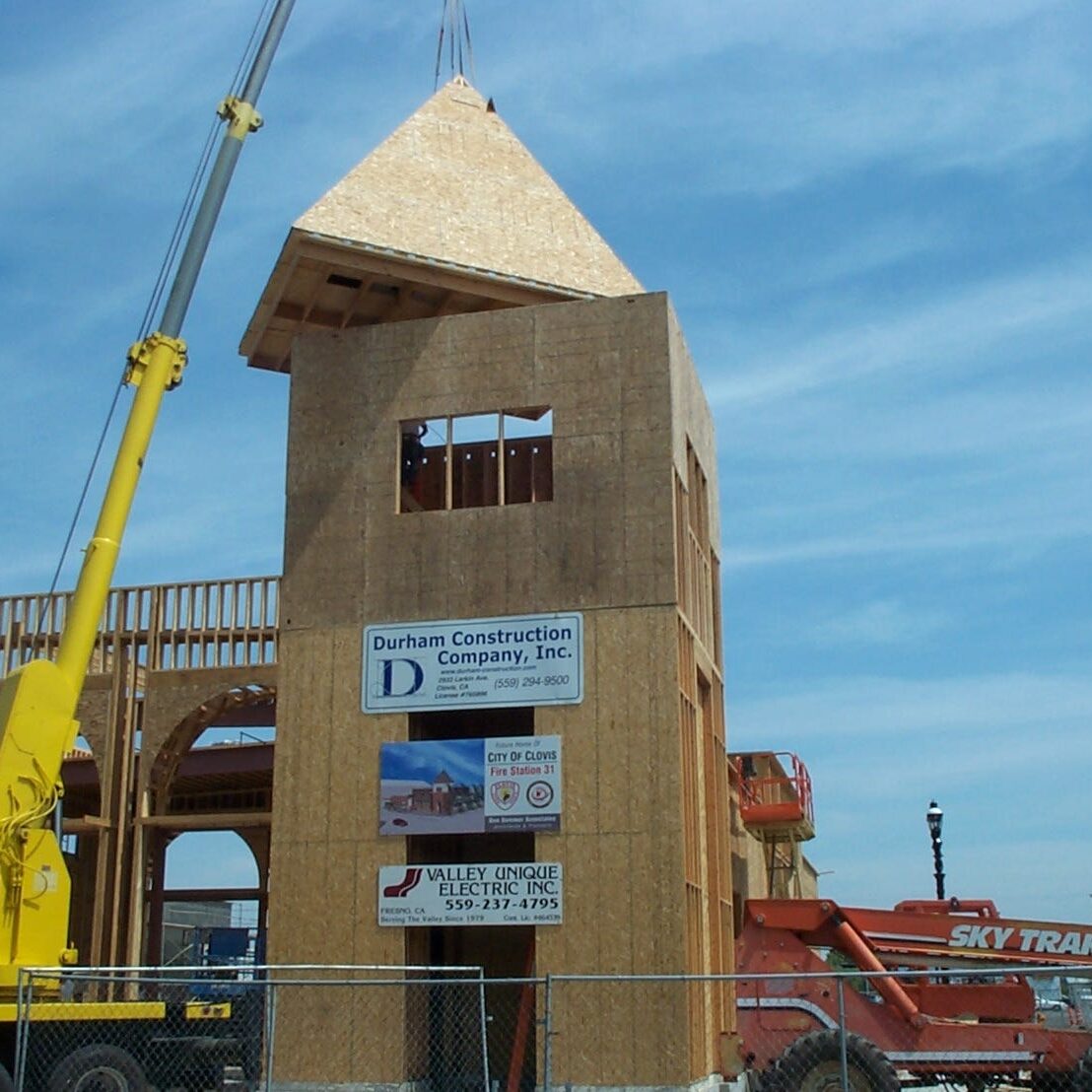 A wide shot of the fire station's tower under construction. A crane is lowering the pyramid-shaped roof onto the top of the plywood-framed tower. A large sign for Durham Construction Company is attached to the side of the building.