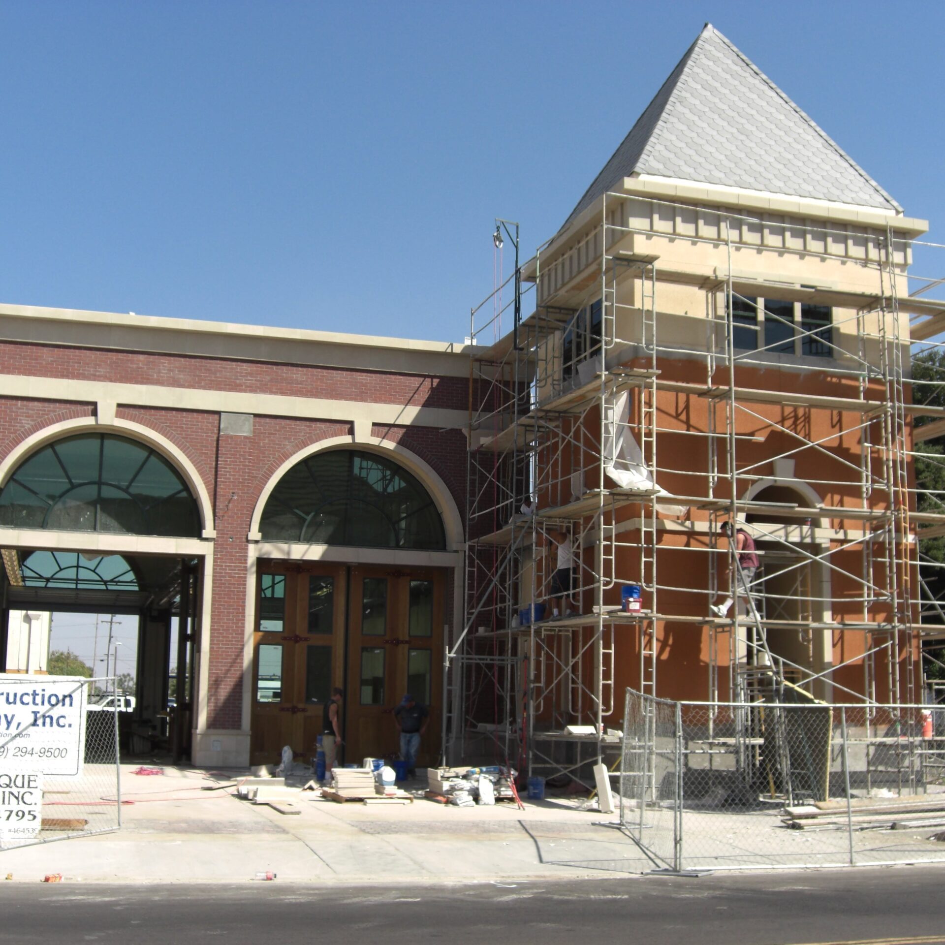 The fire station's exterior nearing completion. Scaffolding is set up around the tower, where workers are applying stucco. The front of the building shows finished brickwork and arched wooden doorways.