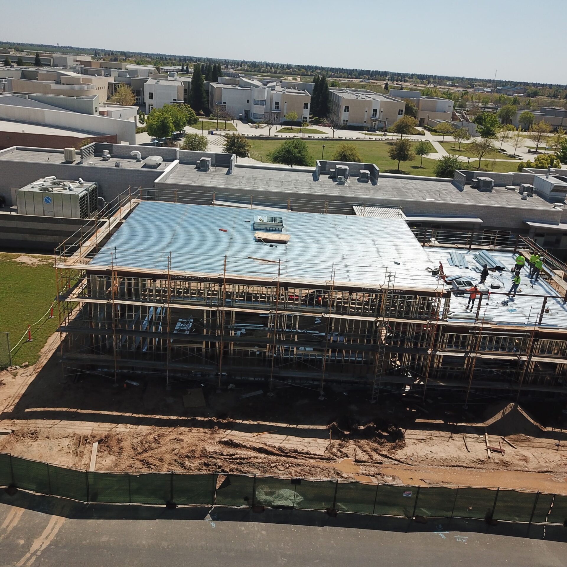An aerial view of a building under construction. The structure is supported by scaffolding, and construction workers are on the roof. The surrounding area includes other school buildings and a grassy field.