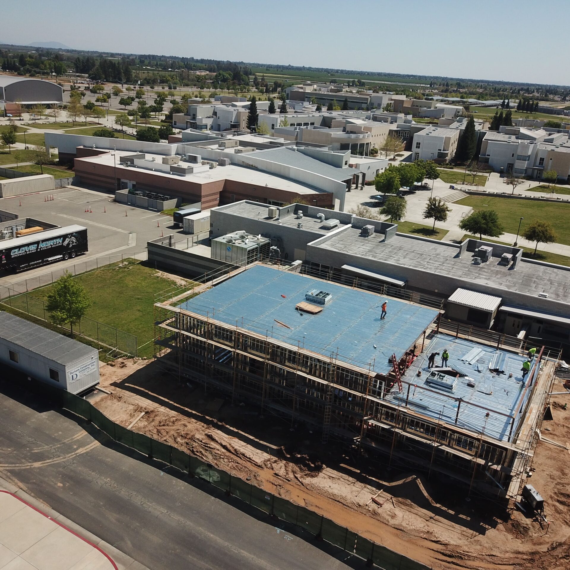 An aerial view of a building under construction, showing a blue roof tarp. Workers are visible on the roof. The building is surrounded by scaffolding and dirt, with other school buildings and a parking lot in the background.