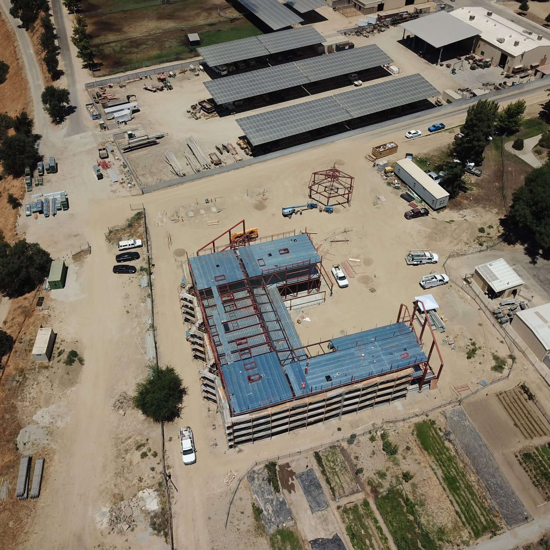 An aerial drone shot of a construction site. The red steel frame of a new building, forming an L-shape, is being erected on a large dirt lot. Various construction vehicles and materials are scattered around the site, with a large, flat building and solar panels in the background.