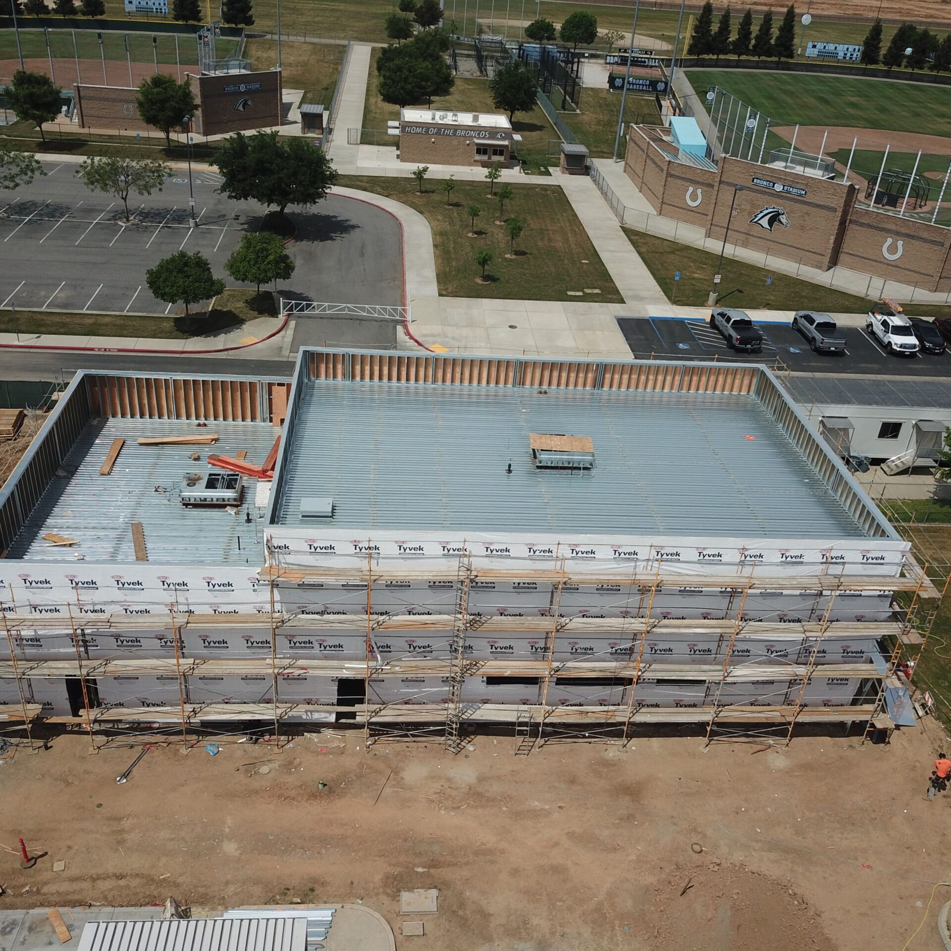 An aerial view of a building under construction. The structure is framed with scaffolding, and the roof is made of corrugated metal. In the background, there is a baseball field and other completed school buildings and parking lots.