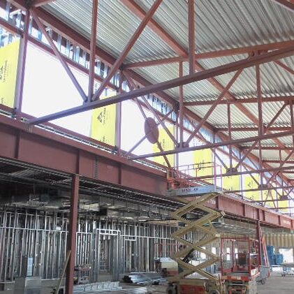 The interior of a building under construction shows exposed red iron trusses supporting the roof. Large rectangular openings in the upper part of the walls are covered with yellow sheathing. Below, metal studs frame the interior walls, and a scissor lift is visible on the concrete floor.