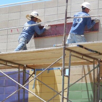Two construction workers, one wearing a straw hat and the other a white hard hat, are on scaffolding. They are installing a rectangular object on a concrete block wall. Below the scaffolding, the wall is covered in a colorful mosaic of purple, yellow, and green tiles.