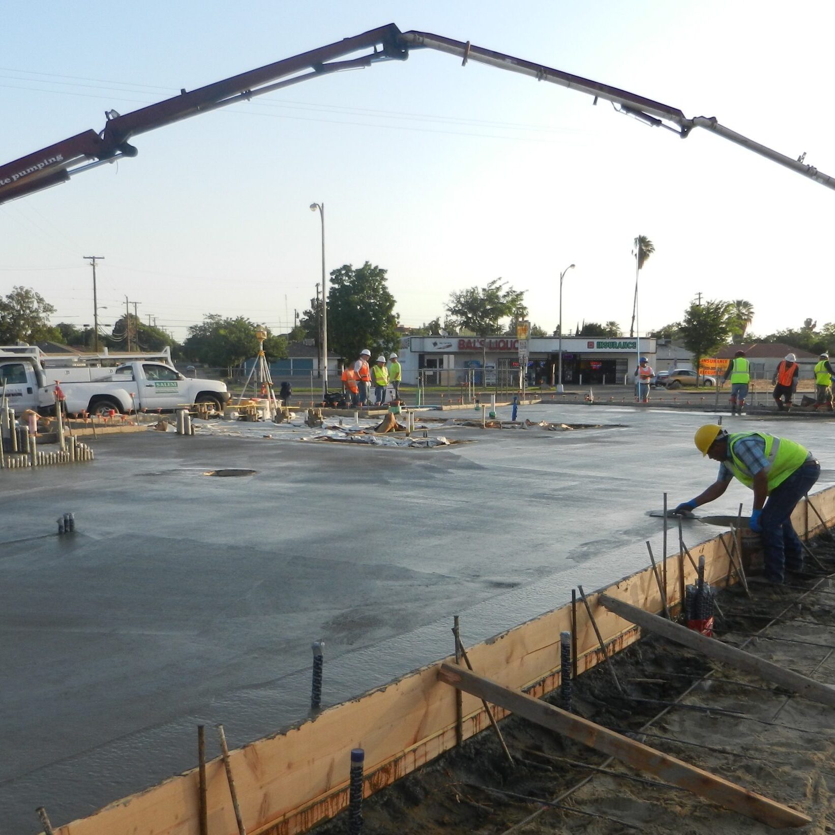 A concrete pump truck with a long boom, labeled "Allied Concrete Pumping," is pouring a large concrete slab. Several construction workers in hard hats and safety vests are on the job site, with some guiding the concrete and another finishing the edges of the formwork. In the background, there are streetlights and a commercial building.