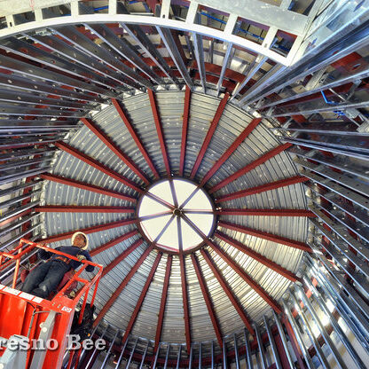 A high-angle view from inside a building under construction shows the circular, domed ceiling framing. A worker in a hard hat is positioned on a red scissor lift, looking up at the intricate network of curved and radial metal beams and studs. The central skylight is visible at the top of the dome. A watermark for "The Fresno Bee" is in the bottom left corner.
