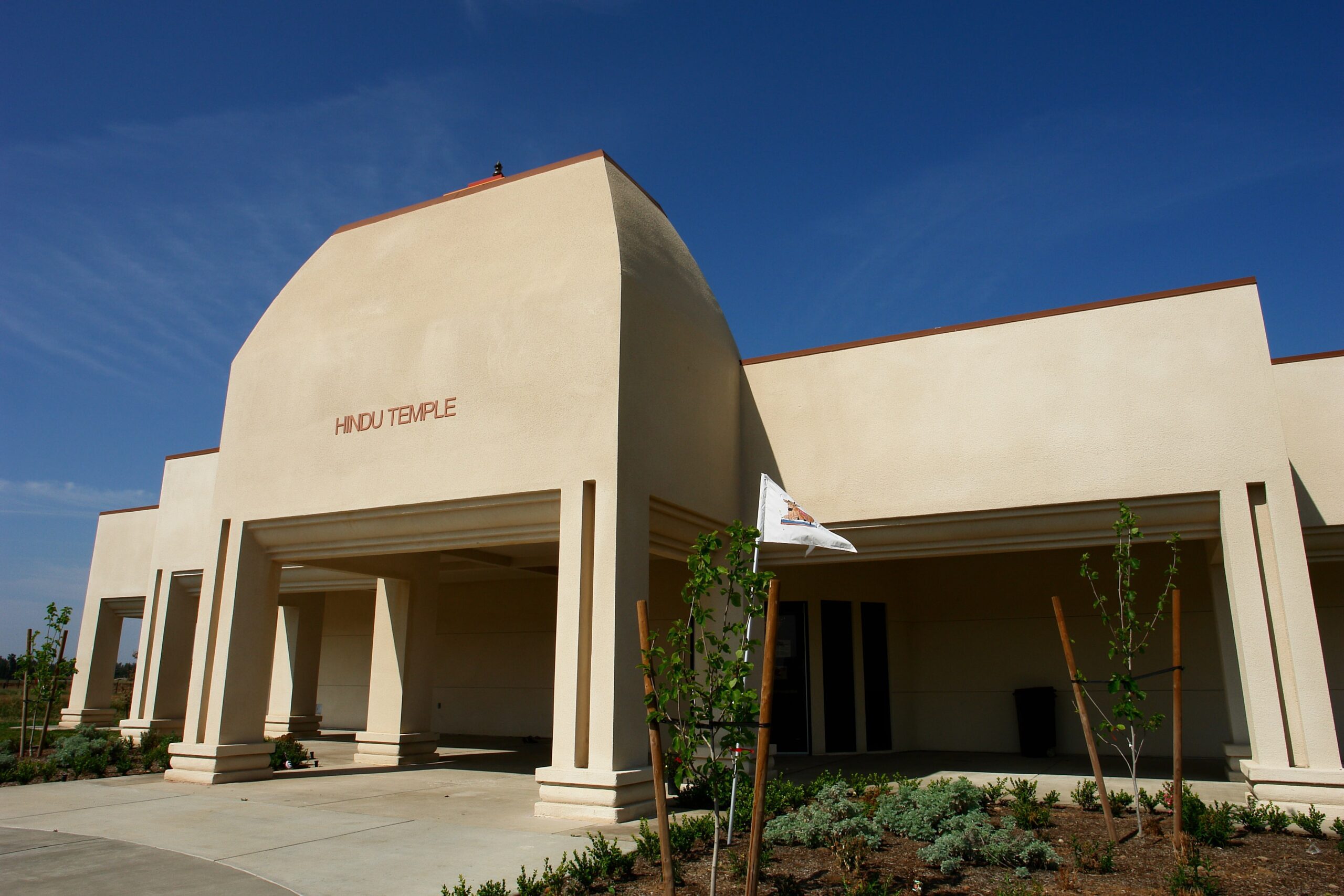Hindu Temple of Clovis A low-angle, exterior shot of a modern Hindu temple building. The structure is one story and has a clean, minimalist design with smooth, light-colored stucco walls. The words "HINDU TEMPLE" are visible in simple lettering on the front facade. The building features an arched entryway and several square columns. The landscaping in the foreground consists of small plants and mulch. A small flag on a pole is visible near the entrance. The sky is a clear, bright blue with a few wispy clouds. The image conveys a sense of peaceful simplicity.
