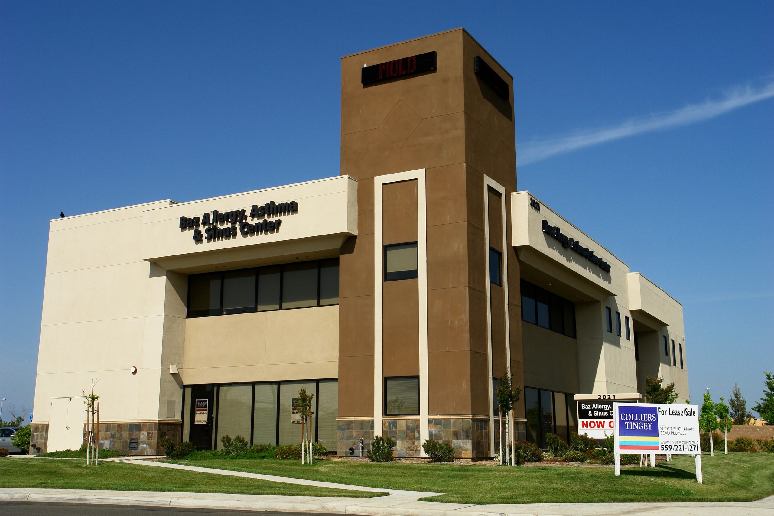 DSC00812-min A slightly low-angle, exterior shot of a modern, two-story commercial building on a sunny day. The building's facade is a mix of light-colored stucco, a darker tan central tower, and a stone veneer base. The words "Ear, Allergy, & Sinus Center" are visible on the left side of the building. In the foreground, there is a manicured lawn and two "For Lease/Sale" signs for Colliers Tingey. The sky is a clear, deep blue. The image captures the building from a street-level perspective, highlighting its clean lines and professional appearance.