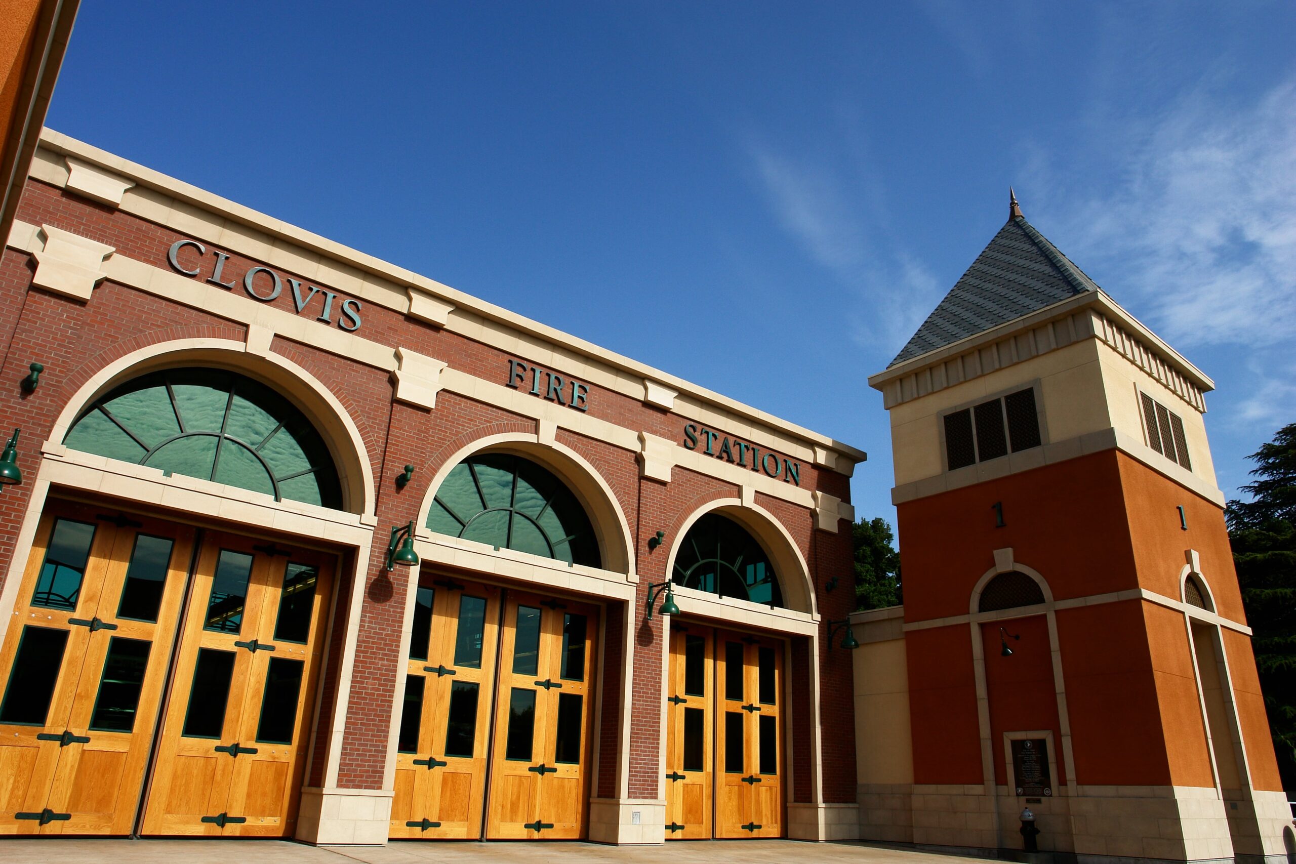 Clovis Fire Station A low-angle, exterior shot of the Clovis Fire Station #1, captured from a perspective that highlights the building's height. The image focuses on the three large arched doors for the fire engines and the prominent tower to the right. The facade is a mix of red brick and orange-red stucco. The words "CLOVIS FIRE STATION" are visible in blue lettering above the arched doors. The doors themselves are made of light-colored wood with numerous small glass panes. The tower has a pointed, gray roof. The sky is a clear, bright blue with a few scattered clouds.