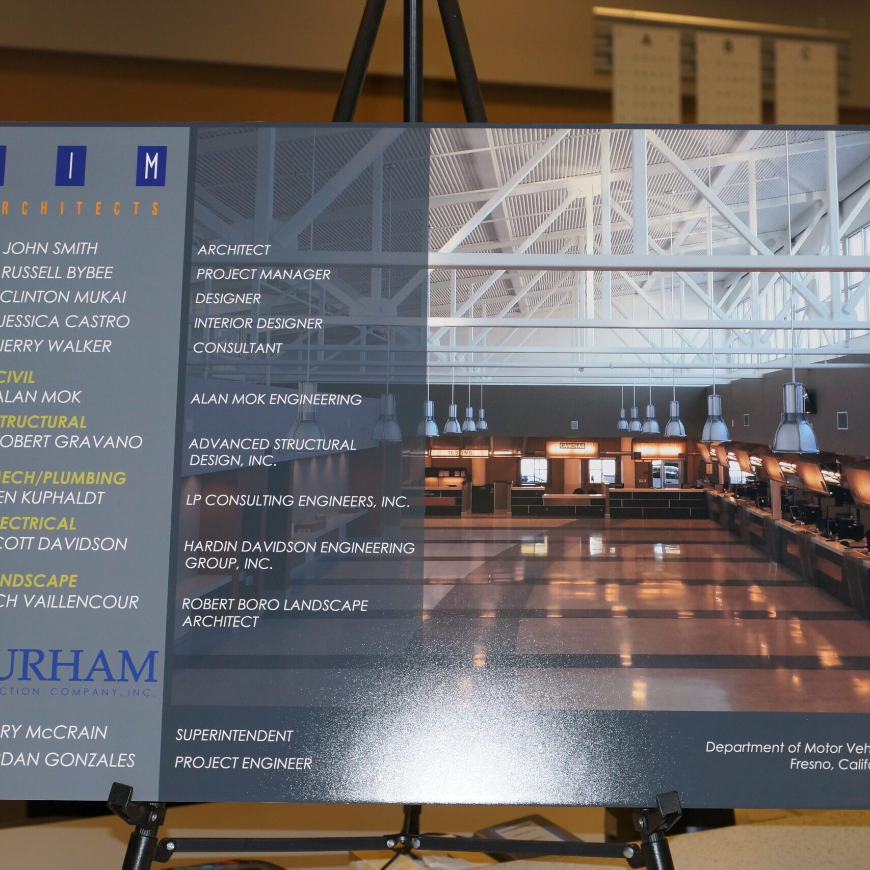 A display board shows a digital rendering of the interior of a DMV office, featuring a spacious room with numerous service counters, modern lighting, and an open, high ceiling with exposed trusses. The left side of the board lists the key professionals and firms involved in the project, including SIM Architects, Durham Construction, and engineers for civil, structural, mechanical, plumbing, electrical, and landscape design.