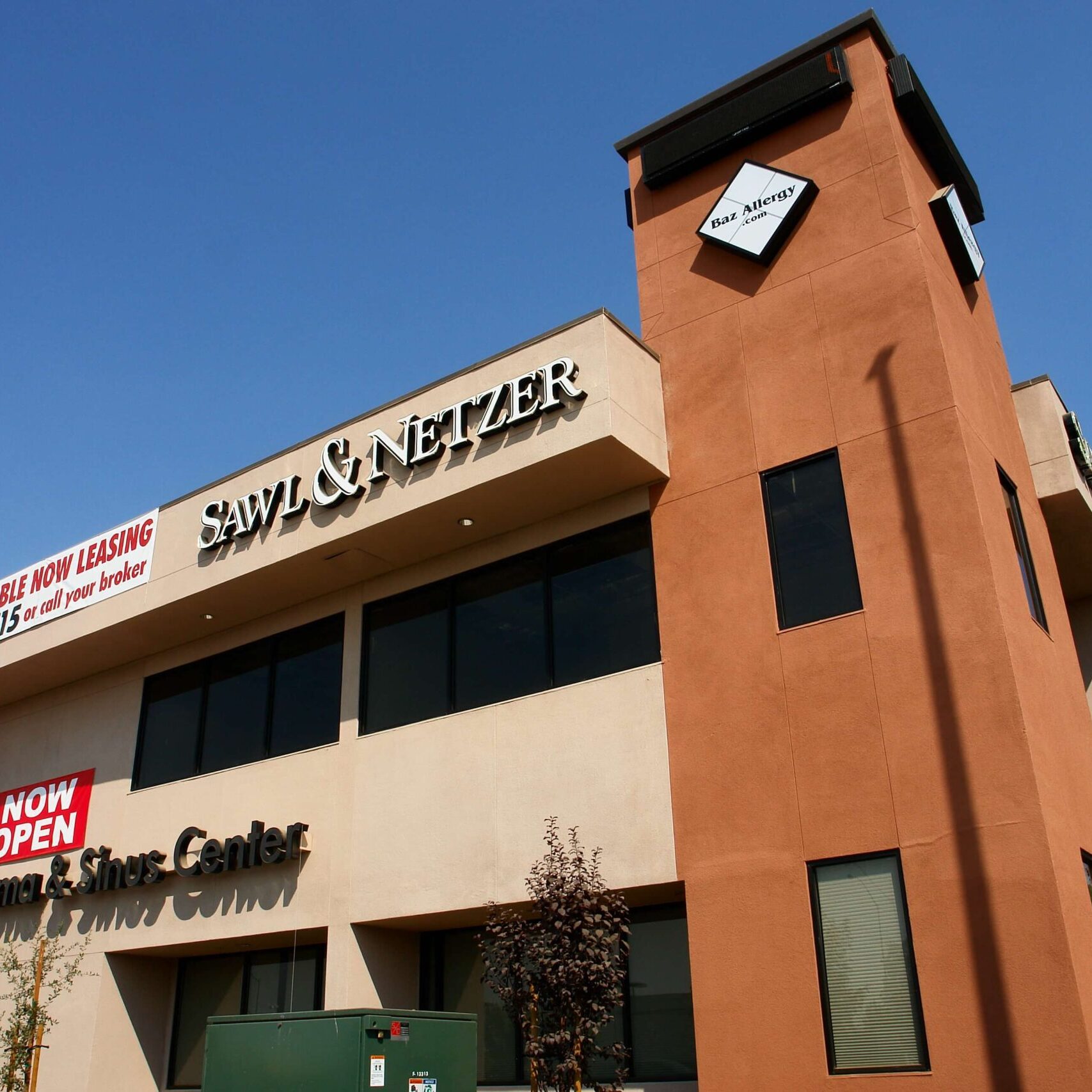 A low-angle shot of a multi-story medical office building with signs for "Sawl & Netzer" and "Baz Allergy, Asthma, & Sinus Center." The building has a prominent brown tower on the right side with a "Baz Allergy" logo. Red banners for "Now Leasing" and "Now Open" are also displayed.