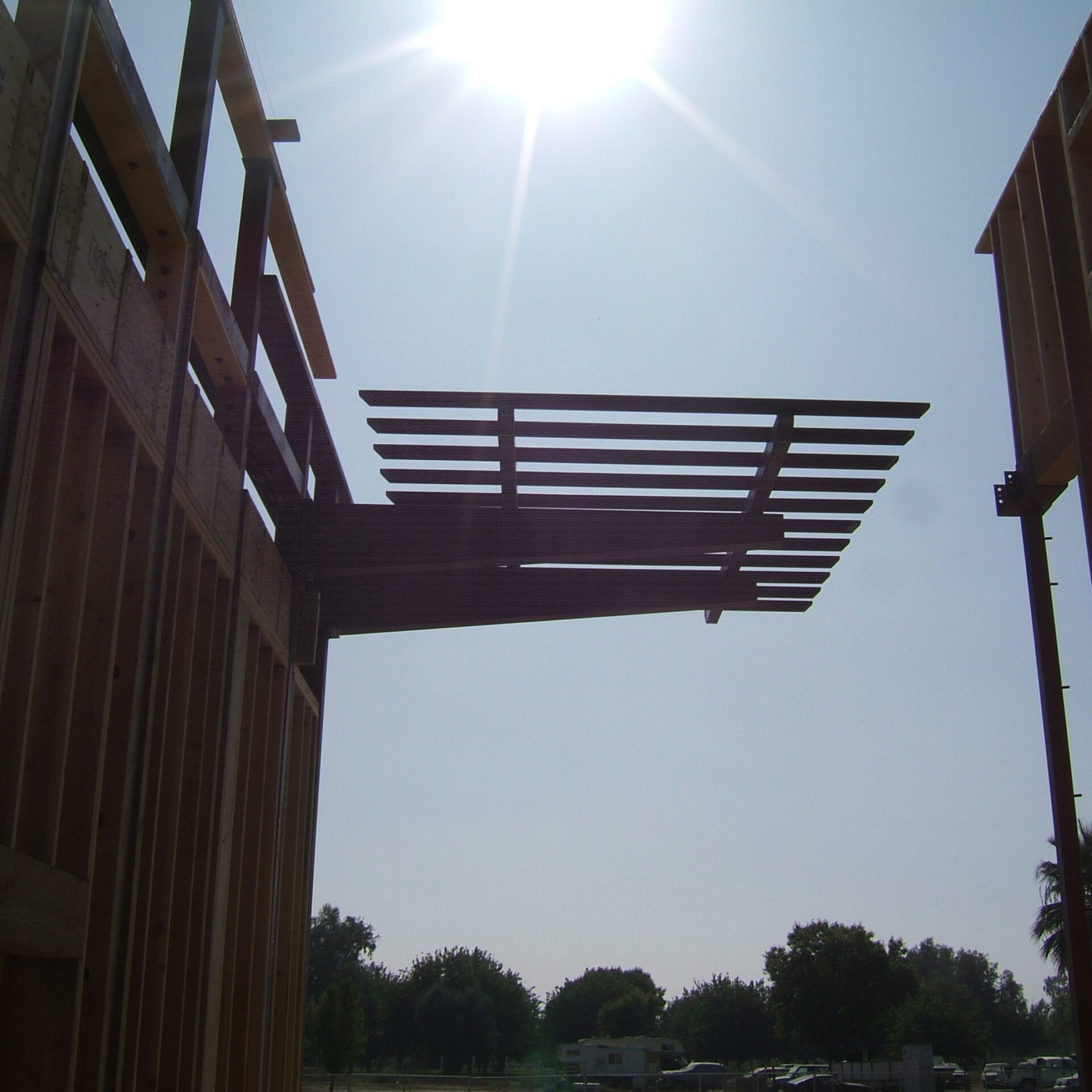 A photo of a building under construction, showing a wooden frame against a bright blue sky. The sun is shining brightly, and there are trees and other vehicles in the background.