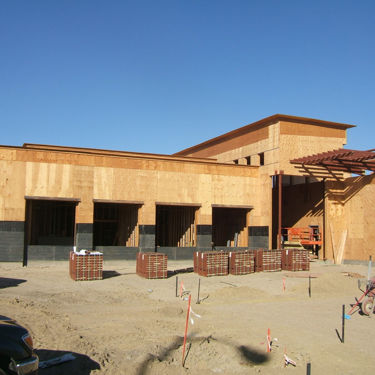 An exterior shot of a building under construction on a sunny day. The walls are a wooden frame covered in plywood. Multiple large rectangular window openings are visible. Construction materials are scattered on the ground, and a truck is parked to the left.