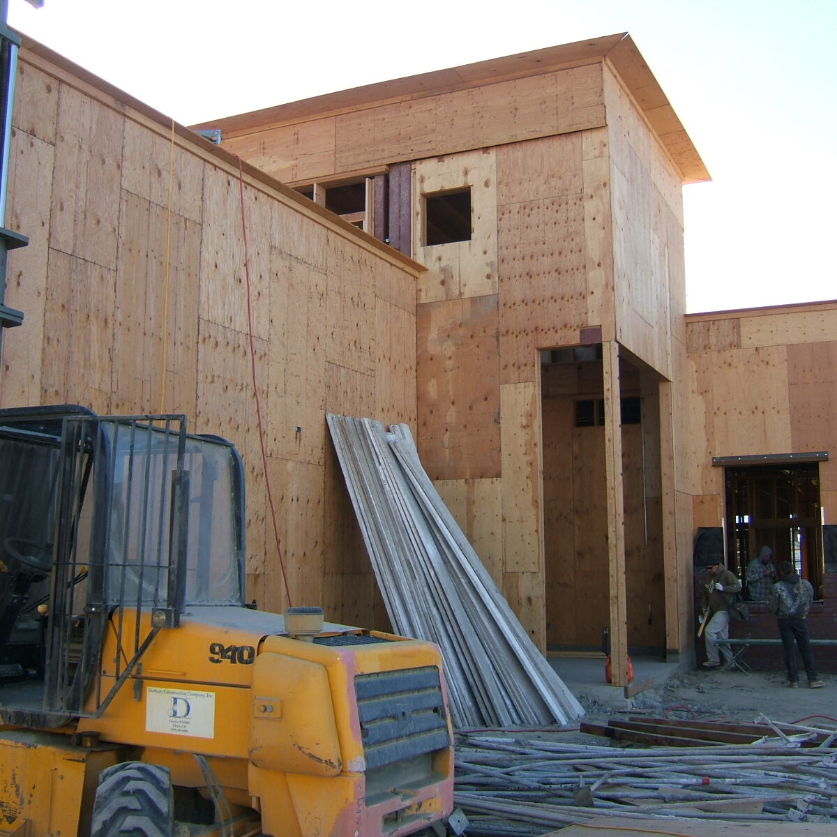 An exterior view of a building under construction. The building is framed with wood and sheathed in plywood. A large yellow forklift is in the foreground, and construction materials are scattered on the dirt ground. People are visible near the building's entrance in the distance.