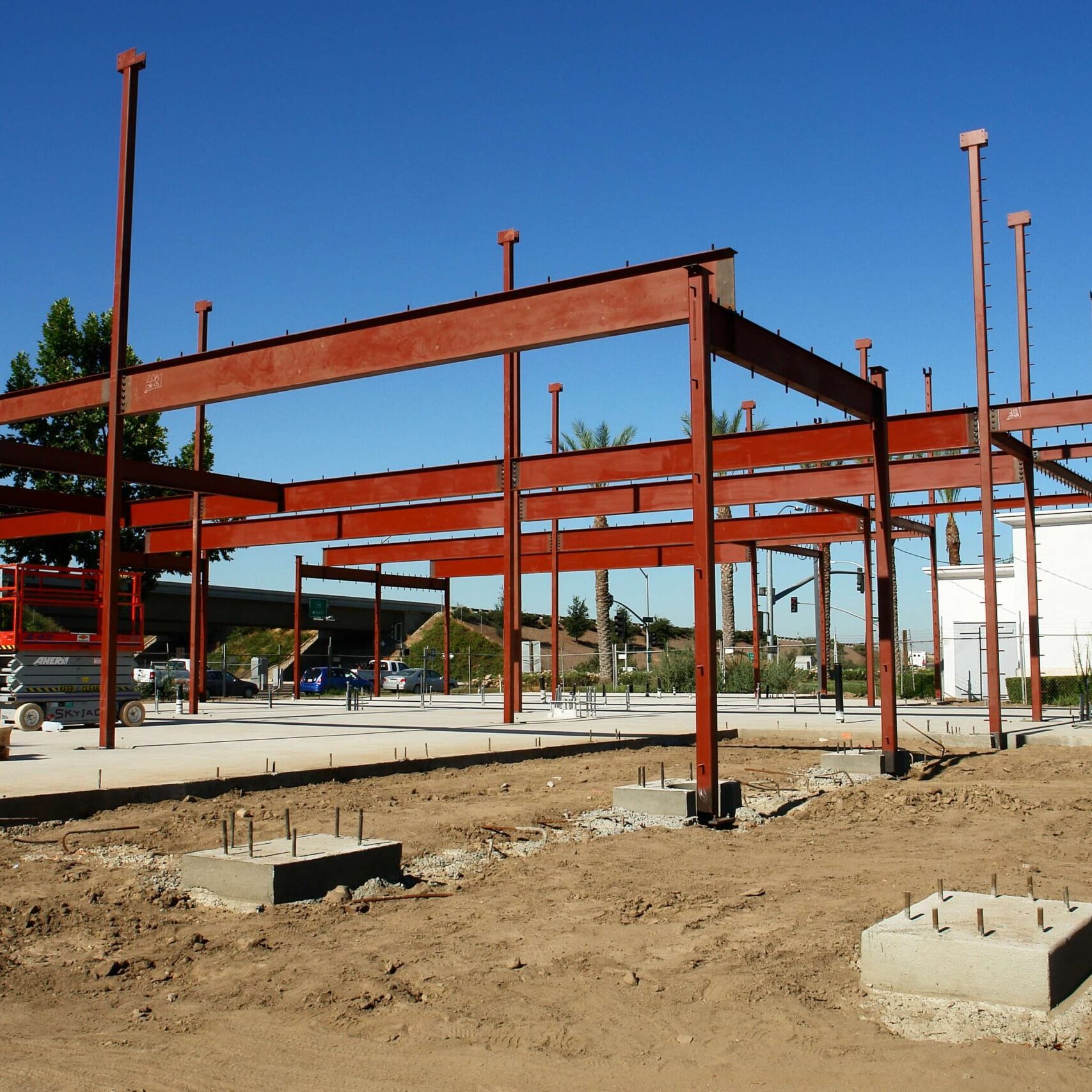 An image of a building under construction. The red steel frame is erected on a dirt lot with concrete footings. The sky is clear and blue.