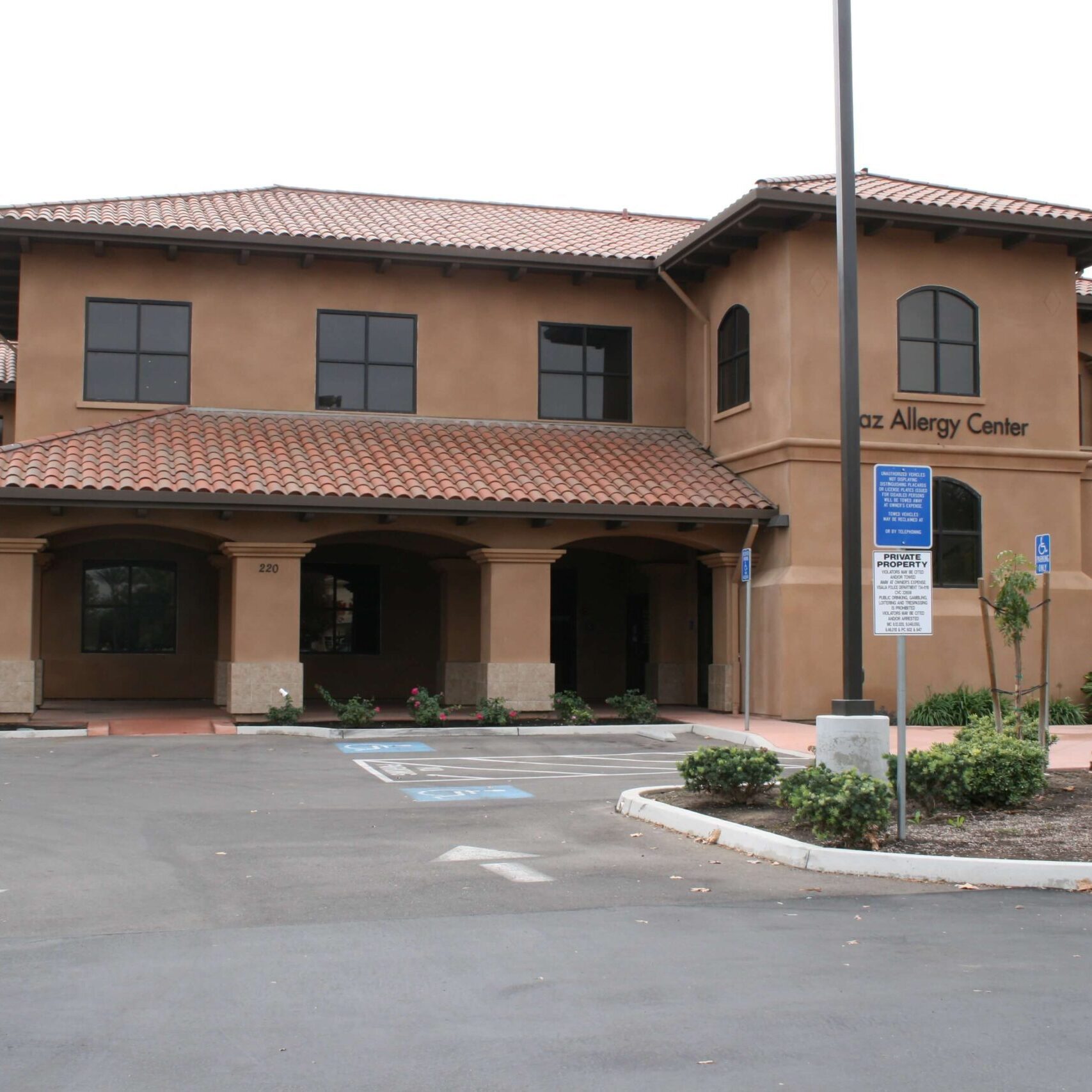 A two-story medical office building in Visalia, California, featuring a Mediterranean-style design with a tile roof and arches. The building is tan and a sign for "Baz Allergy Center" is visible on the right side.