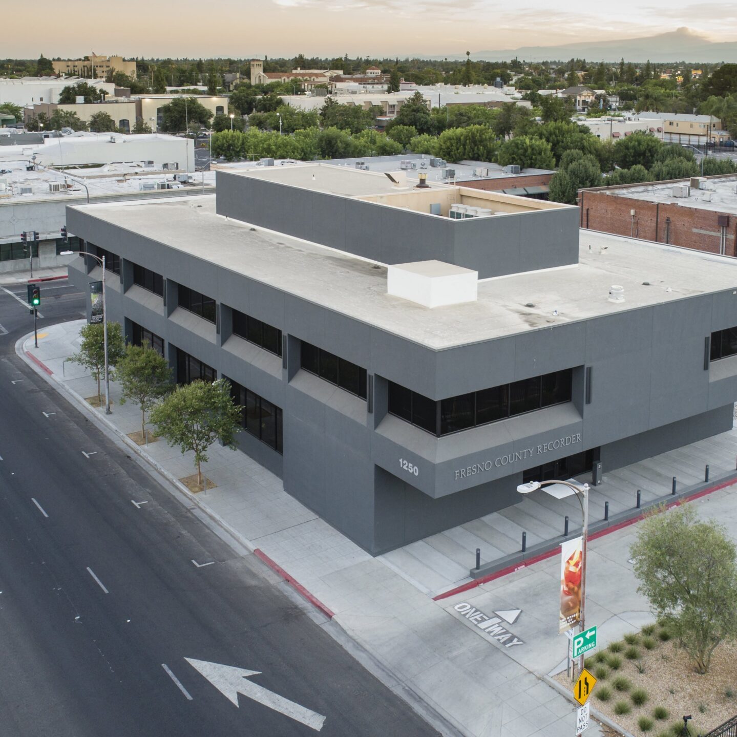 An aerial shot of a modern, two-story, gray building with large, dark, horizontal windows. The building is on the corner of a street intersection with a "One Way" sign pointing toward the building. The words "FRESNO COUNTY RECORDER" are visible on the side. The surrounding area includes several low-rise buildings with flat roofs and a street with traffic lights and arrows painted on the asphalt. Trees and green spaces are scattered throughout the neighborhood, and a hazy sky is visible in the background.