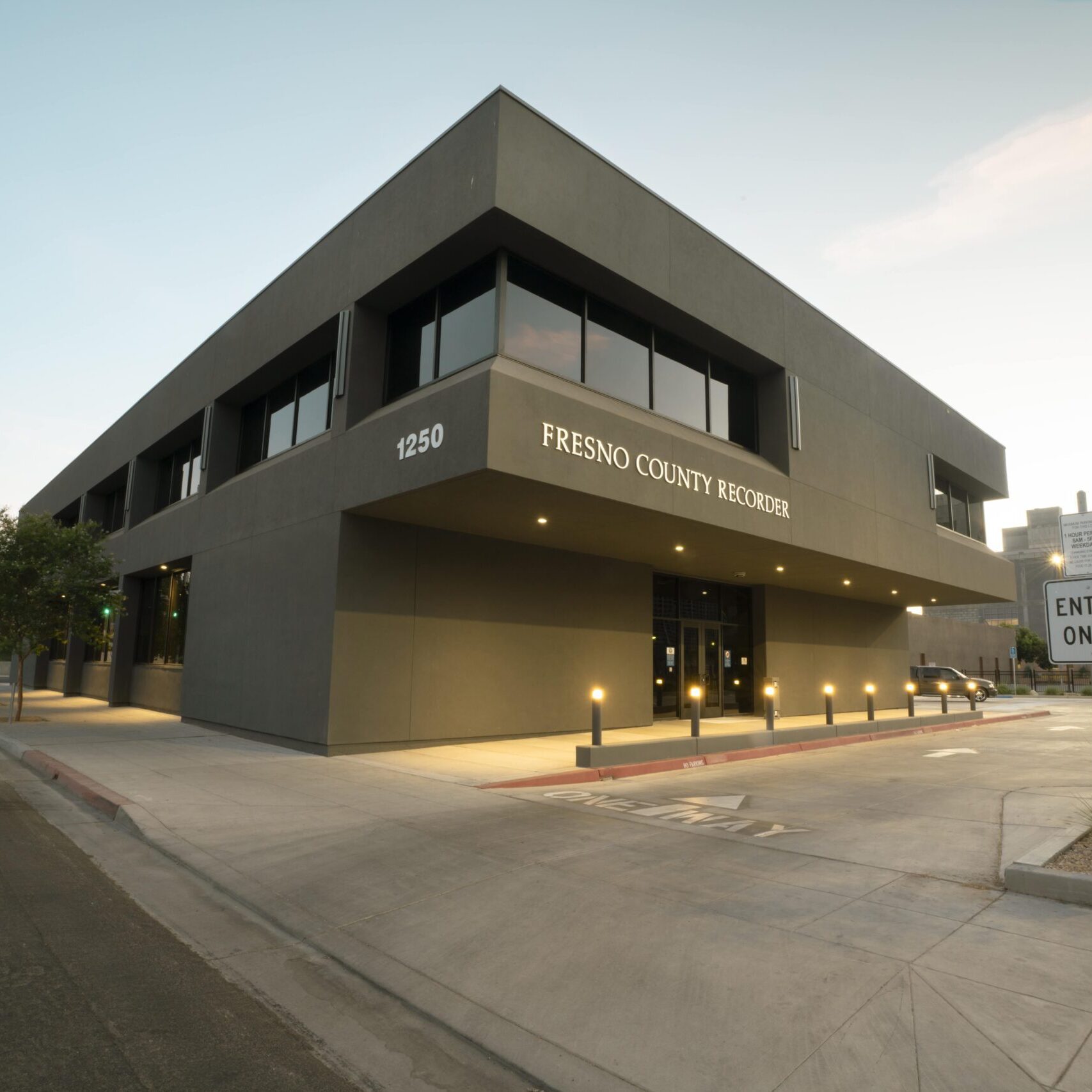 A low-angle, eye-level shot of a modern, two-story, dark gray building with the words "Fresno County Recorder" and "1250" on the facade. The building has large, dark horizontal windows and is lit by several short, glowing lights along the front. A sign that reads "ENTER ONLY" and "PRIVATE PROPERTY" is visible on the right. The sun is setting, and the building is visible from a street corner, with an empty street and sidewalk in the foreground.
