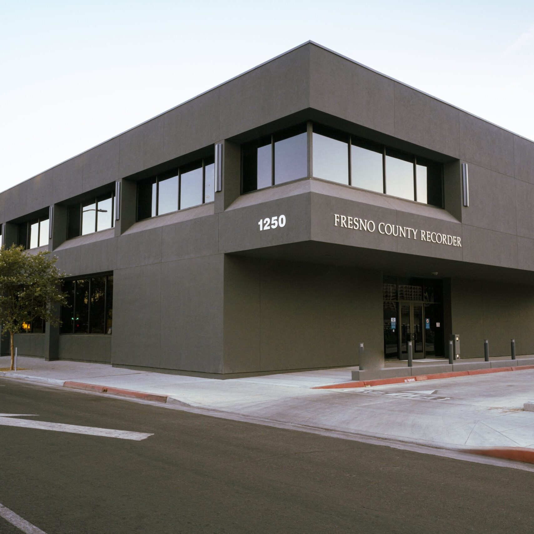 A low-angle, eye-level shot of a modern, two-story, dark gray building with the words "Fresno County Recorder" and "1250" on the facade. The building has large, dark horizontal windows that reflect the sky and the street. An arrow is painted on the empty street, pointing to the building's entrance. A sign that reads "ENTER ONLY" is visible on the right. A small tree and a street light are visible on the left. The sun is setting, and the sky is a pale blue and pink.