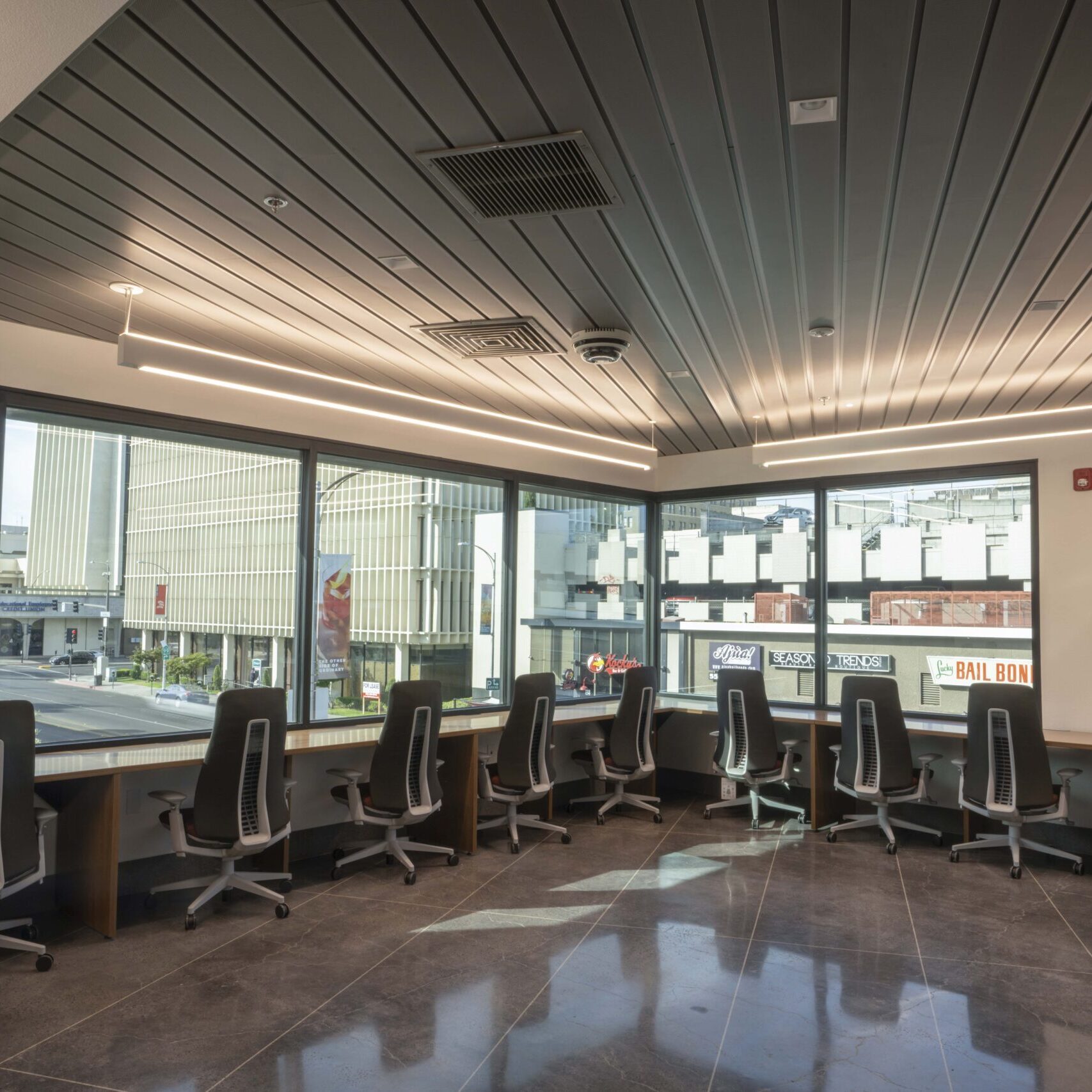 A low-angle, eye-level shot of a modern, empty waiting room or office lobby. The floor is a dark gray, reflective tile. A long, angular customer service counter with gray granite panels and transparent dividers is in the center. The ceiling has a combination of dark gray panels with long recessed lights and a dropped ceiling with square tiles. The walls are plain white.