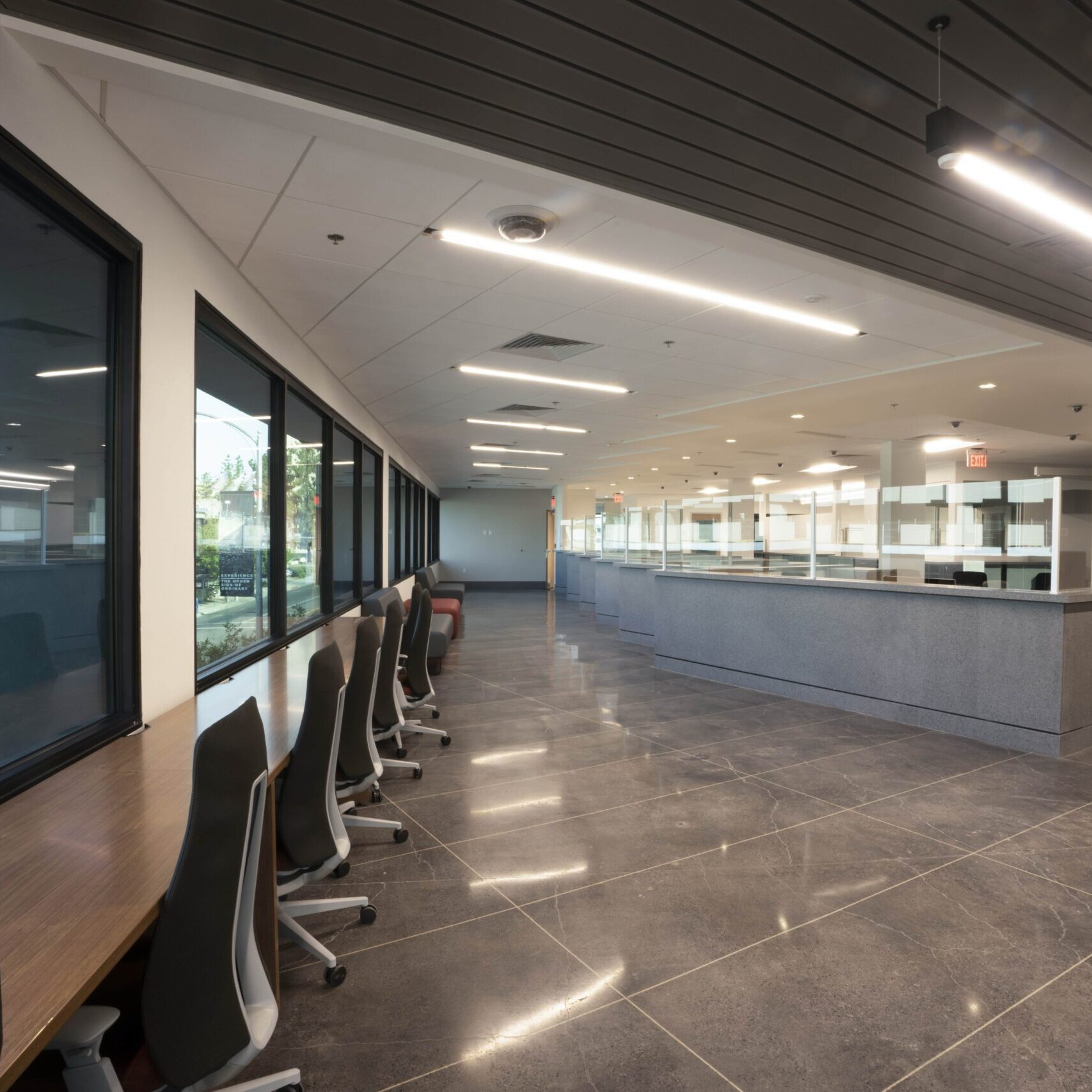 A wide-angle, low-shot of a modern, empty office or waiting area. On the left, a long wooden countertop with a row of rolling black office chairs is visible along a large window that looks out onto the street. Another building with a pointed roof is reflected in the glass. In the background, a long customer service counter with transparent dividers and a gray granite front is visible. The floor is a dark gray, reflective tile.