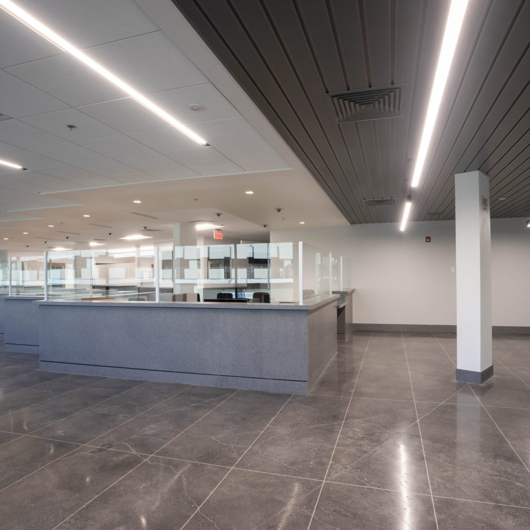 A low-angle, eye-level shot of a modern office or waiting area with a large, angular window that wraps around the room. A row of rolling black office chairs is arranged along a long, low table that follows the curve of the window. The window offers a view of a city street and other buildings. The ceiling is a combination of dark gray panels and dropped tiles with long, bright, recessed lights. The floor is a dark, reflective tile.
