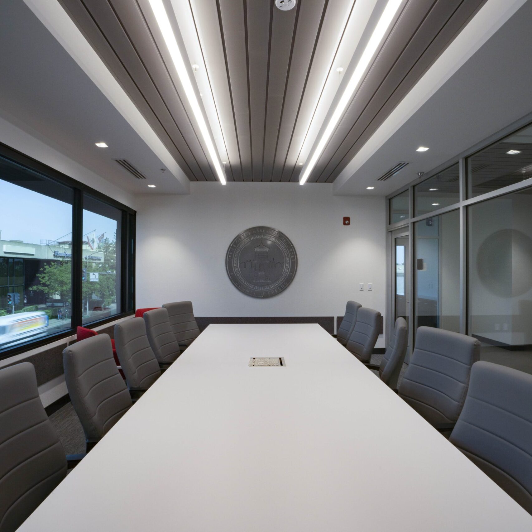 A wide-angle, eye-level shot of a modern conference room. A long, white conference table with several gray rolling chairs is in the center. The walls are white, with a large, gray, circular seal mounted on the back wall. Large windows on the left provide a view of a city street and other buildings. A glass wall on the right looks into another room. The ceiling is a combination of dropped tiles and dark gray panels with long, bright, recessed lights.