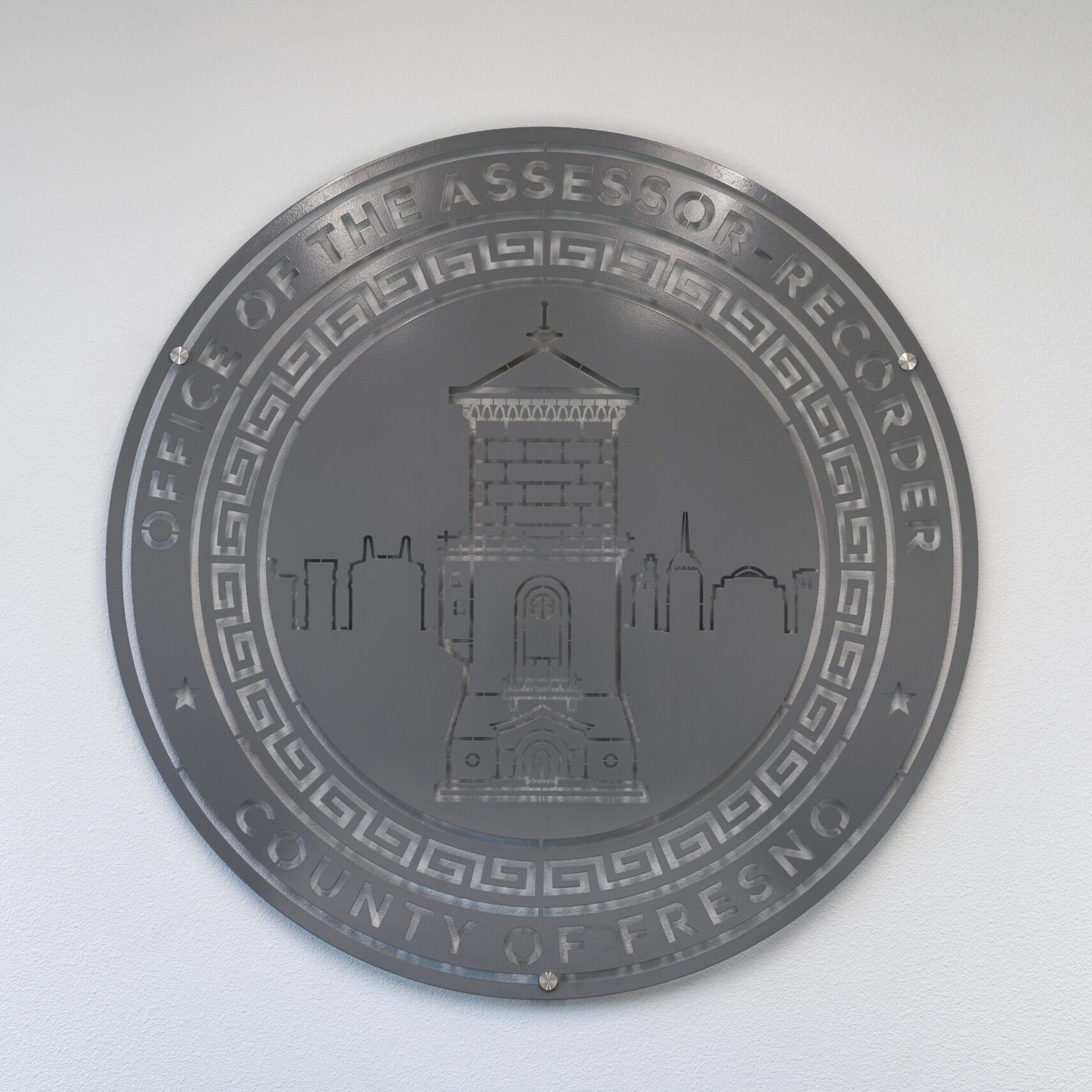 A close-up, eye-level shot of a large, circular emblem etched into a dark gray tile floor. The seal features a drawing of the Fresno Water Tower and the words "OFFICE OF THE ASSESSOR-RECORDER" and "COUNTY OF FRESNO" written around the perimeter. The floor is clean and reflective, and the lines of the tile are visible.