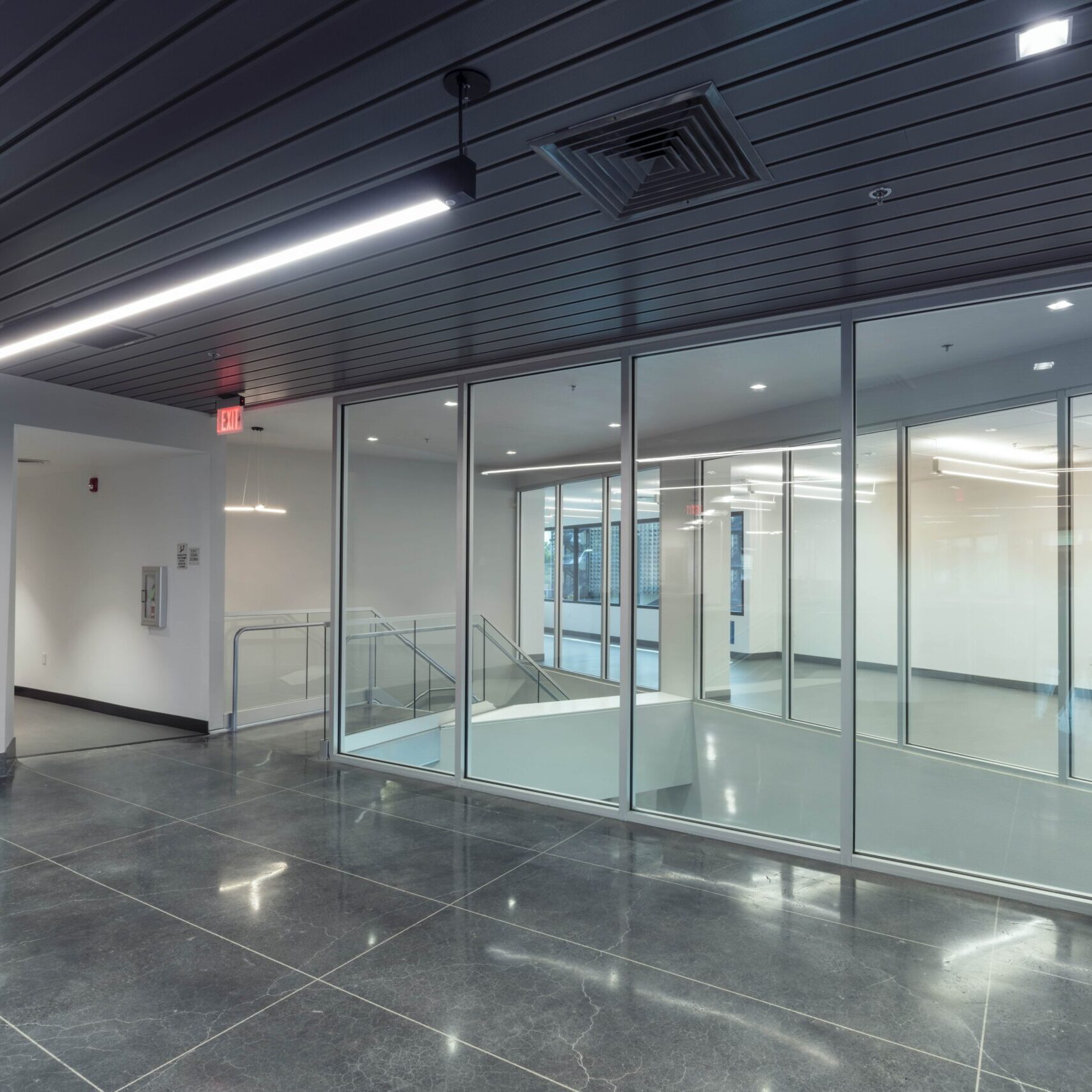 A wide-angle, low-angle shot of a large, modern office lobby. In the foreground, there is a staircase with a gray, steel handrail. In the center, a large, dark gray customer service counter with transparent dividers stretches into the background. The floor is a dark, reflective tile with the Fresno County Assessor-Recorder seal etched into it. The room has high ceilings with several light fixtures and a plain white wall on the right.