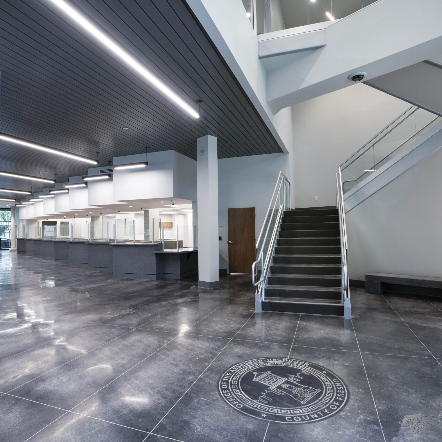 A wide-angle, low-angle shot of a large, modern office lobby. In the foreground, there is a staircase with a gray, steel handrail. In the center, a large, dark gray customer service counter with transparent dividers stretches into the background. The floor is a dark, reflective tile with the Fresno County Assessor-Recorder seal etched into it. The room has high ceilings with several light fixtures and a plain white wall on the right.