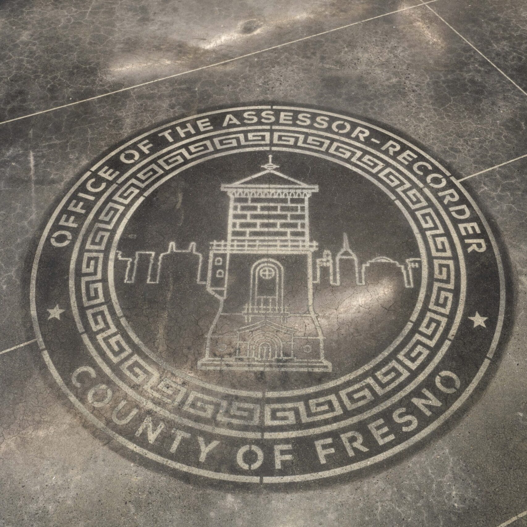 A close-up, eye-level shot of a large, circular emblem etched into a dark gray tile floor. The seal features a drawing of the Fresno Water Tower and the words "OFFICE OF THE ASSESSOR-RECORDER" and "COUNTY OF FRESNO" written around the perimeter. The floor is clean and reflective, and the lines of the tile are visible.