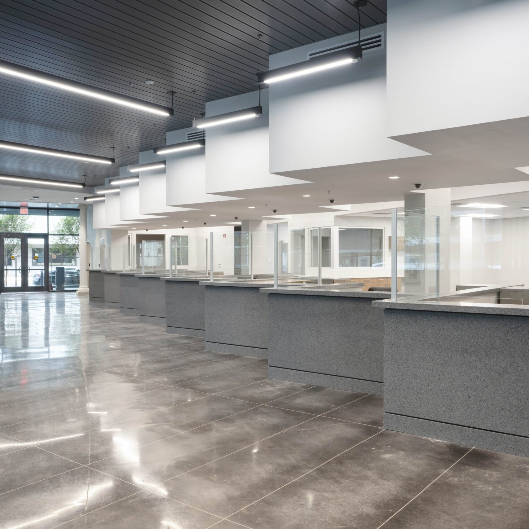 A low-angle, eye-level shot of a long series of customer service counters in a large, modern office. The counters are a dark, speckled gray with transparent dividers. The floor is a dark, reflective tile, and the ceilings are a combination of dark gray panels and dropped tiles with long, bright, recessed lights. Large glass doors are visible at the far end of the room.
