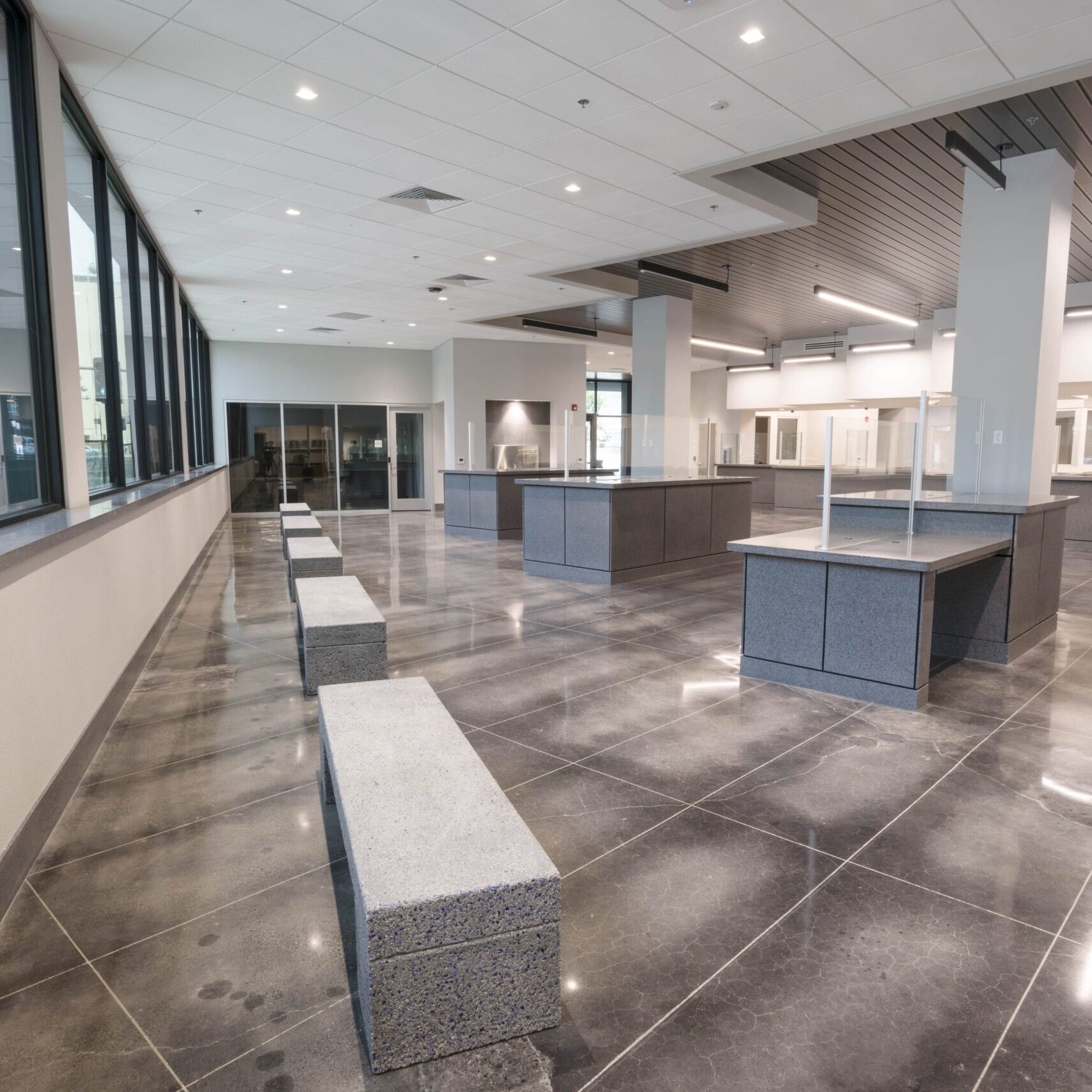 A low-angle, eye-level shot of a modern, empty waiting room or office lobby. A row of long, gray, granite benches is arranged in the foreground. In the background, a series of service counters with transparent dividers is visible. The floor is a dark, reflective tile. A wall of windows on the left provides a view of a street and another building. The room has high ceilings with a combination of dropped tiles and dark gray panels with long, bright, recessed lights.