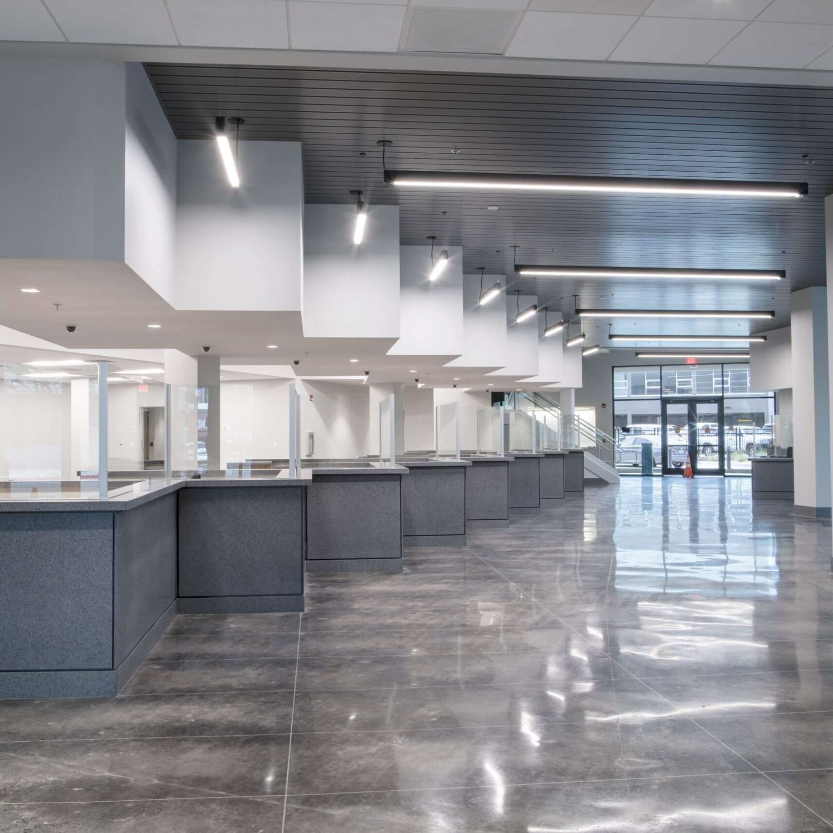 A low-angle, eye-level shot of a long series of customer service counters in a large, modern office. The counters are a dark, speckled gray with transparent dividers. The floor is a dark, reflective tile, and the ceilings are a combination of dark gray panels and dropped tiles with long, bright, recessed lights. A fire extinguisher is mounted on the wall to the left. Large glass doors are visible at the far end of the room.